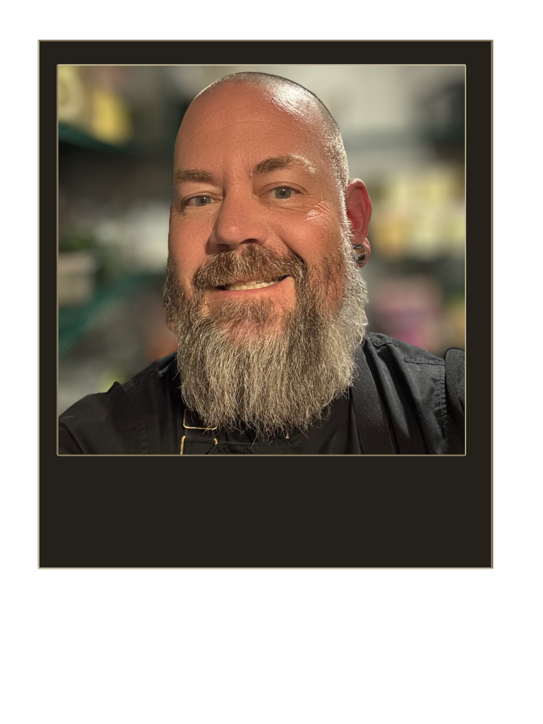 A close-up selfie of a smiling man with a bald head, blue eyes, and a full beard with a mustache. He is wearing a black shirt and earrings, with a blurred background that appears to be an indoor setting, possibly a kitchen.
