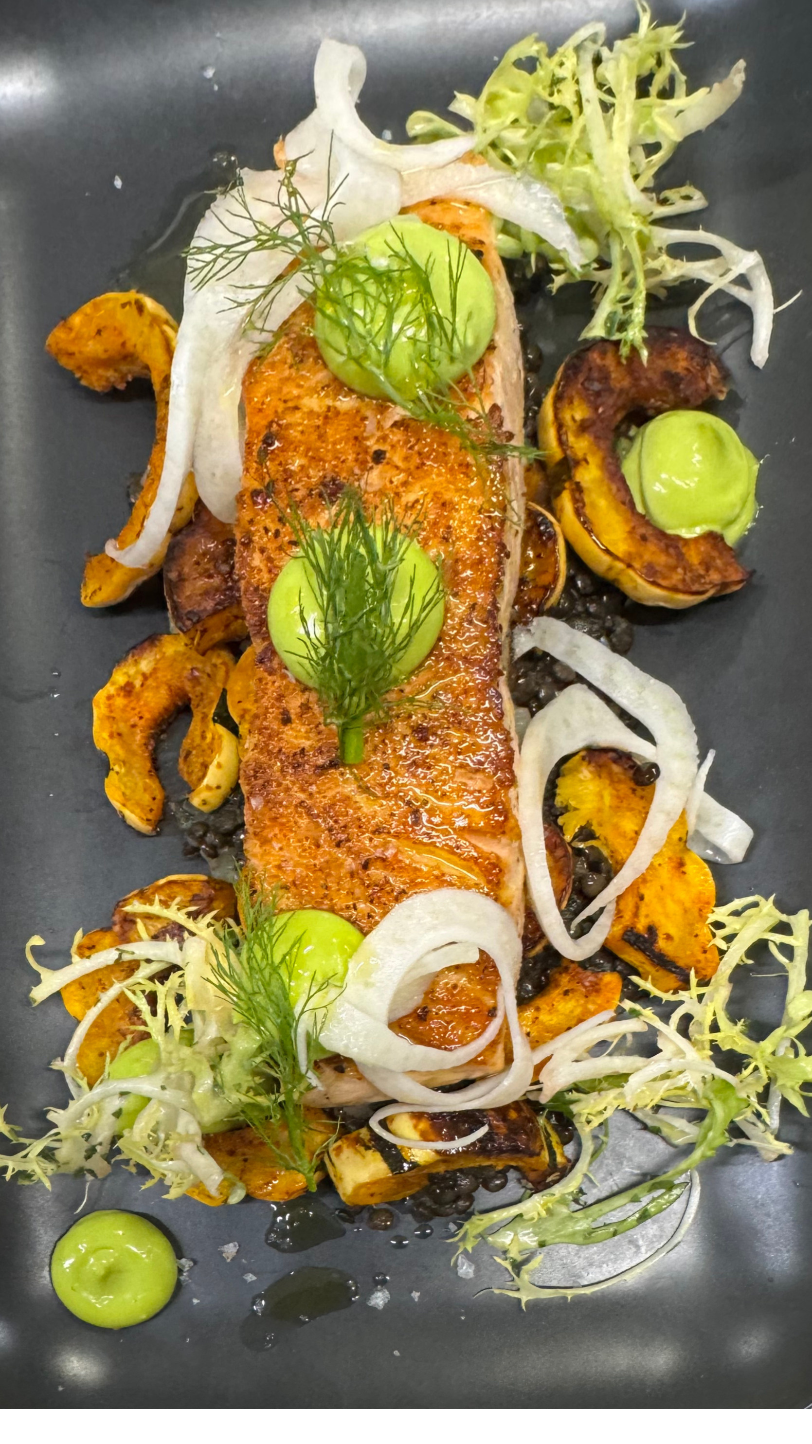 Grilled salmon fillet with charred vegetables, garnished with fresh herbs, onion slices, and dots of green sauce on a black plate.