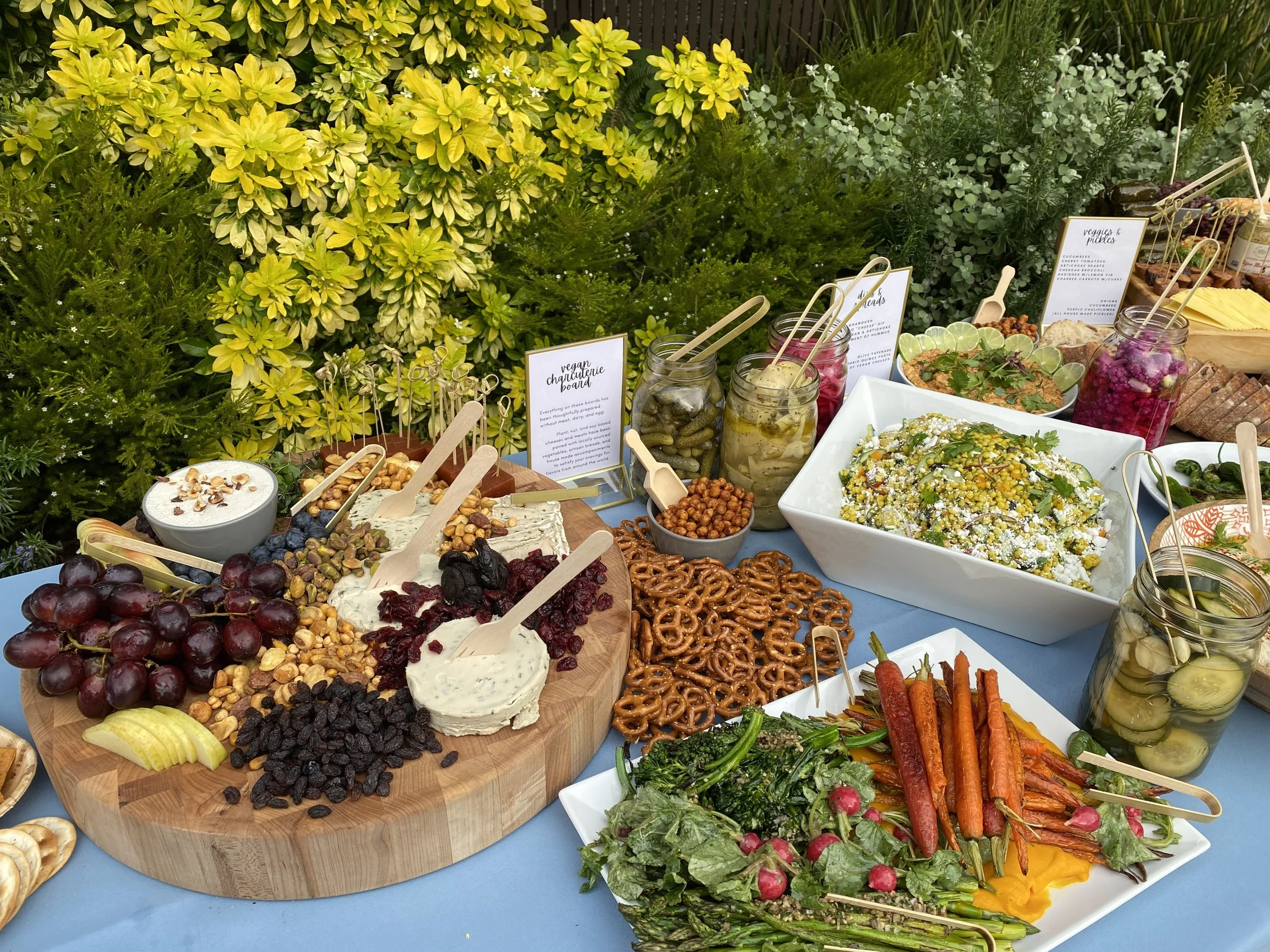 A colorful spread of healthy foods including grapes, dried fruits, veggie and dip platter, salads, pickles, and vegetables with greenery and signs for dietary labels on a table.