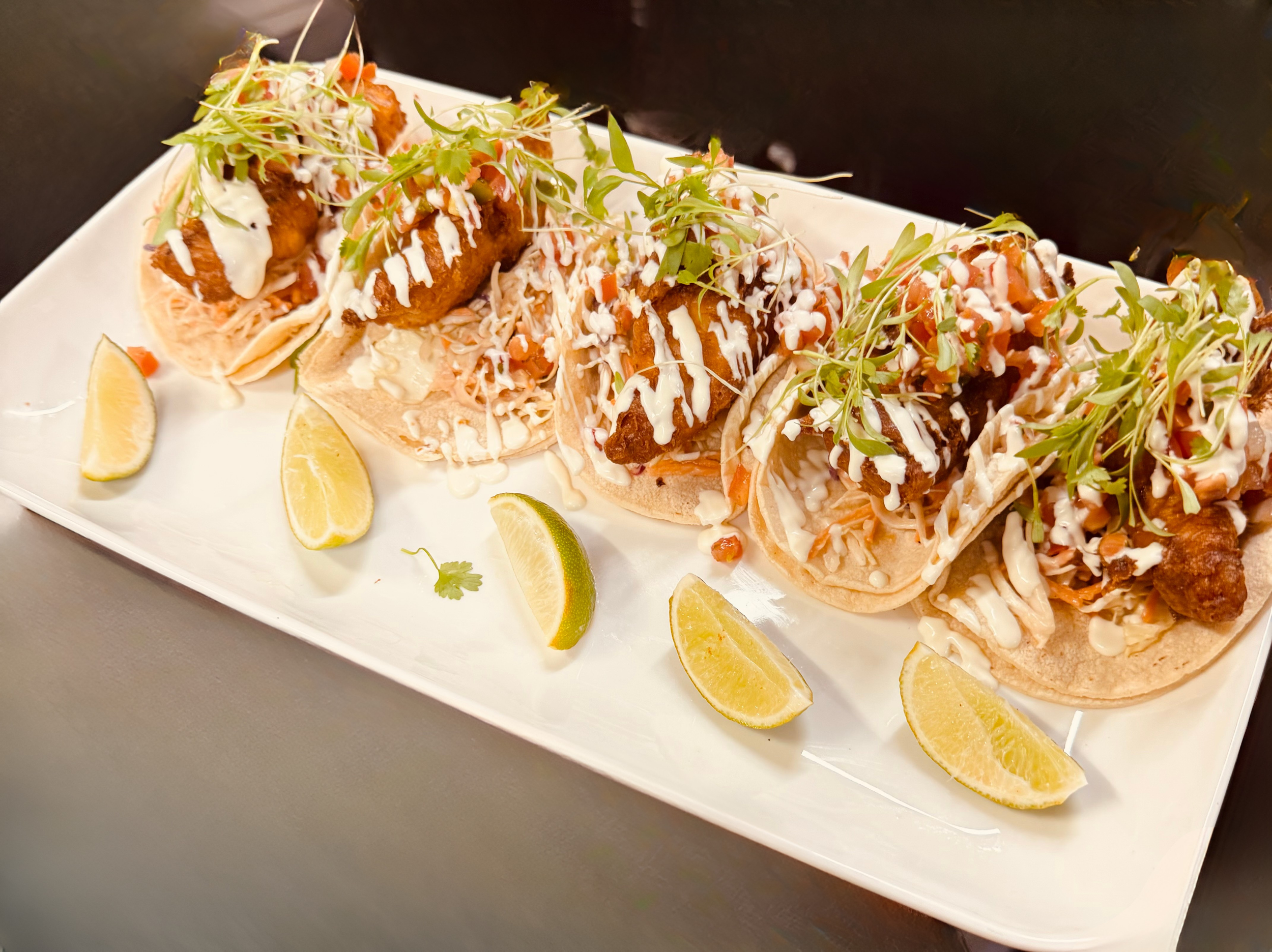 Close-up of a white rectangular plate with four fish tacos topped with microgreens and drizzled with white sauce, garnished with lemon wedges around the plate.