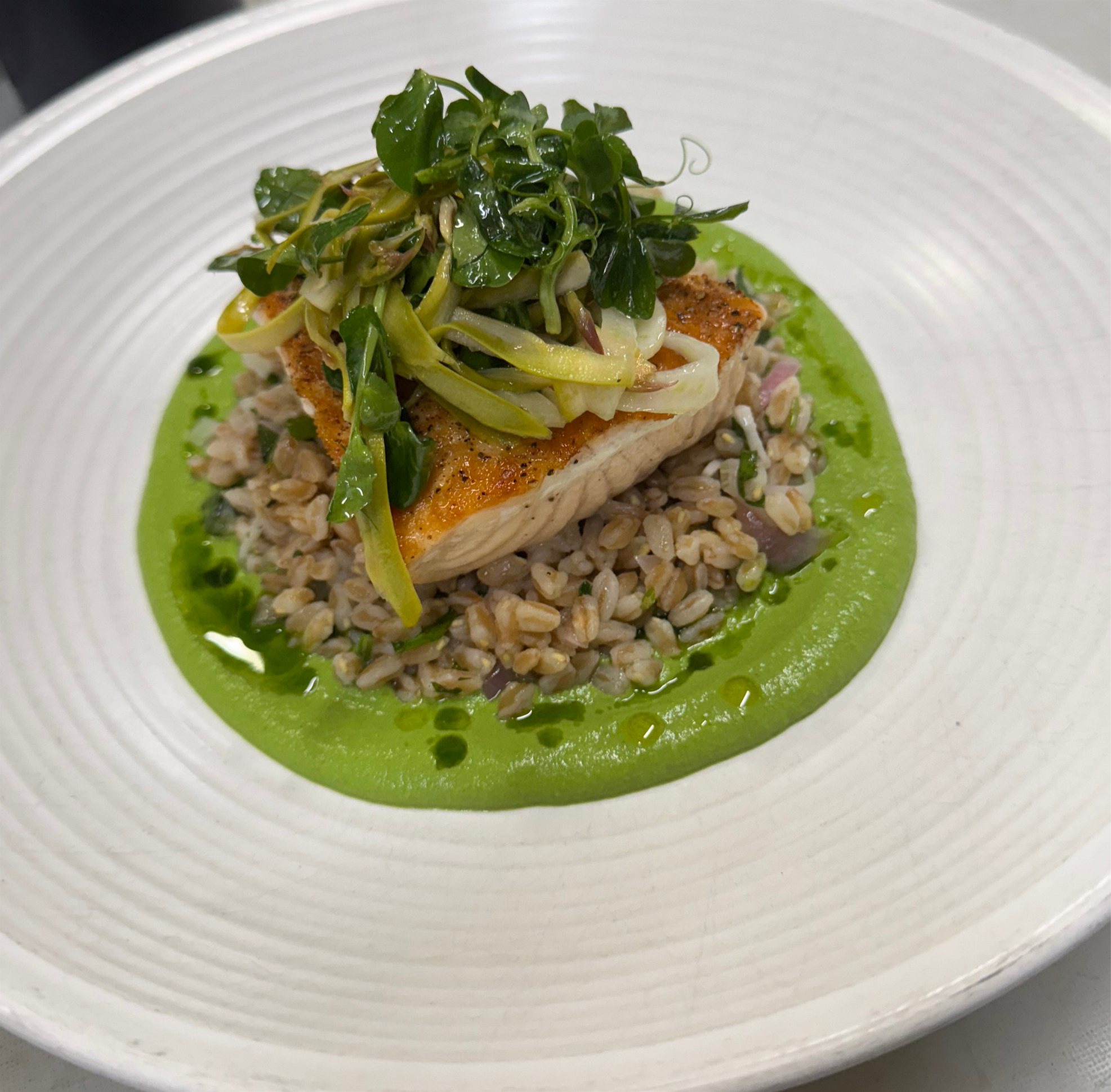 A plated dish featuring a piece of cooked fish topped with sautéed green vegetables and served on a bed of rice, with a green sauce or puree surrounding the rice.