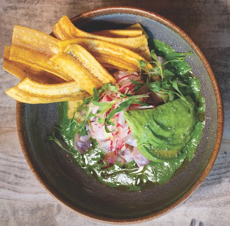 Tuna poke bowl with sliced avocado, radish, microgreens, and fried plantains in a bowl with green sauce.