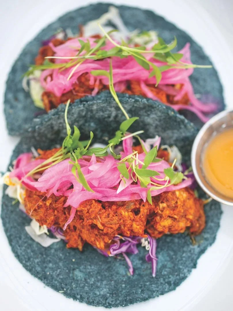 Two blue corn tacos filled with fried chicken, topped with pickled pink onions and microgreens, served with a side of yellow sauce.