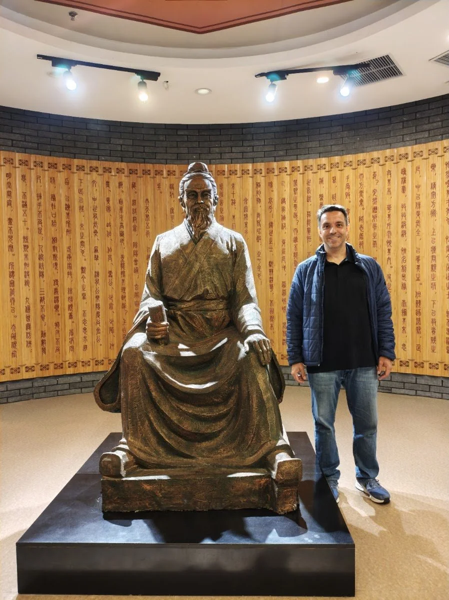 Homem posando ao lado de uma estátua de um sábio antigo da Medicina Chinesa, com fundo de painéis de madeira com caracteres chineses.