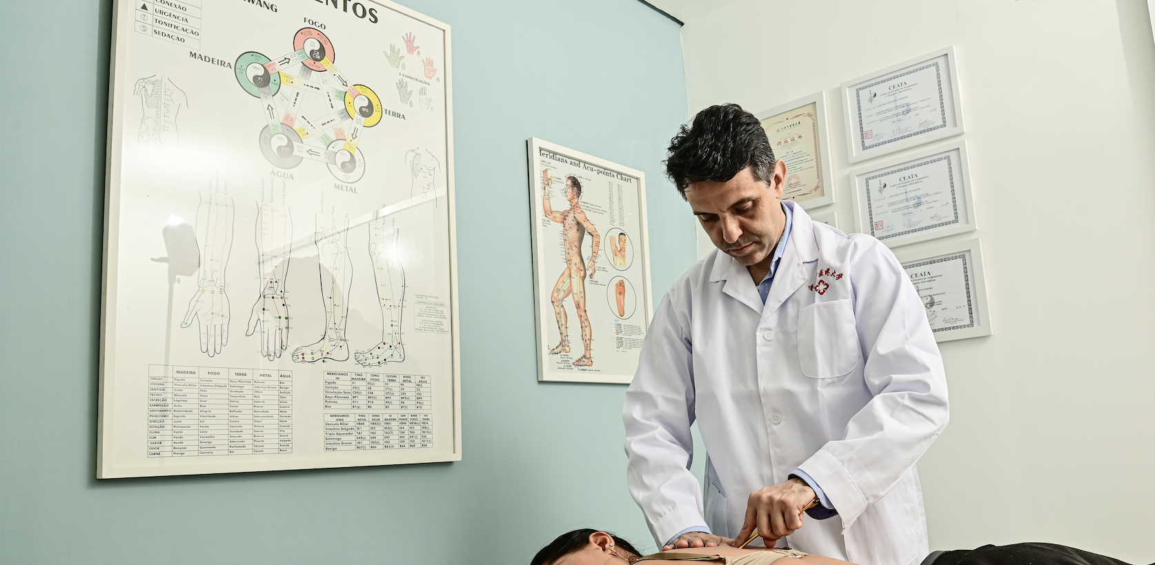 Profissional realizando acupuntura em paciente deitado em uma maca, com quadros de anatomia e certificados na parede ao fundo.