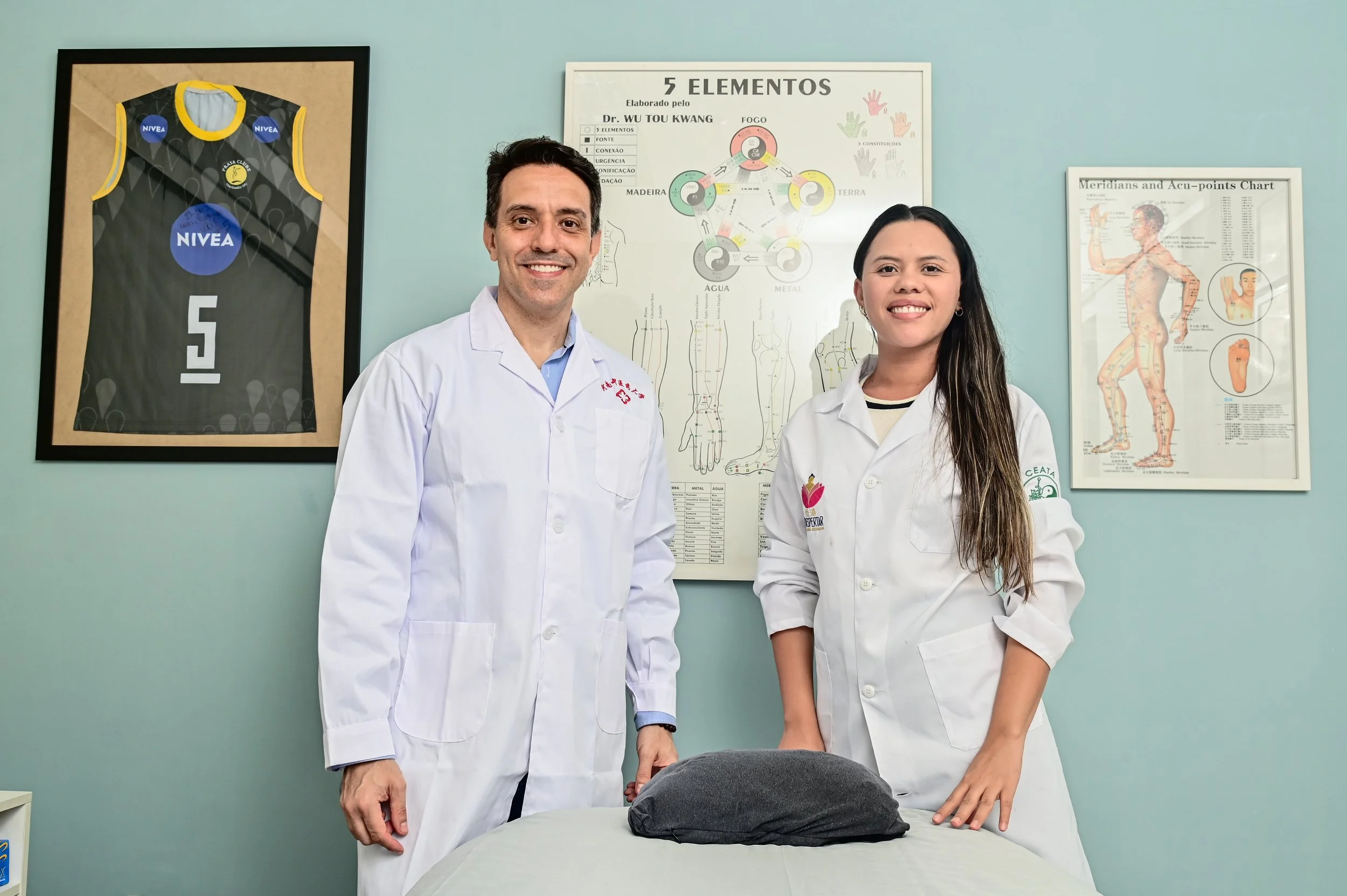 Dois profissionais de jaleco estão em uma sala, um homem e uma mulher, sorrindo para a câmera, com posters de anatomia e elementos de saúde nas paredes.