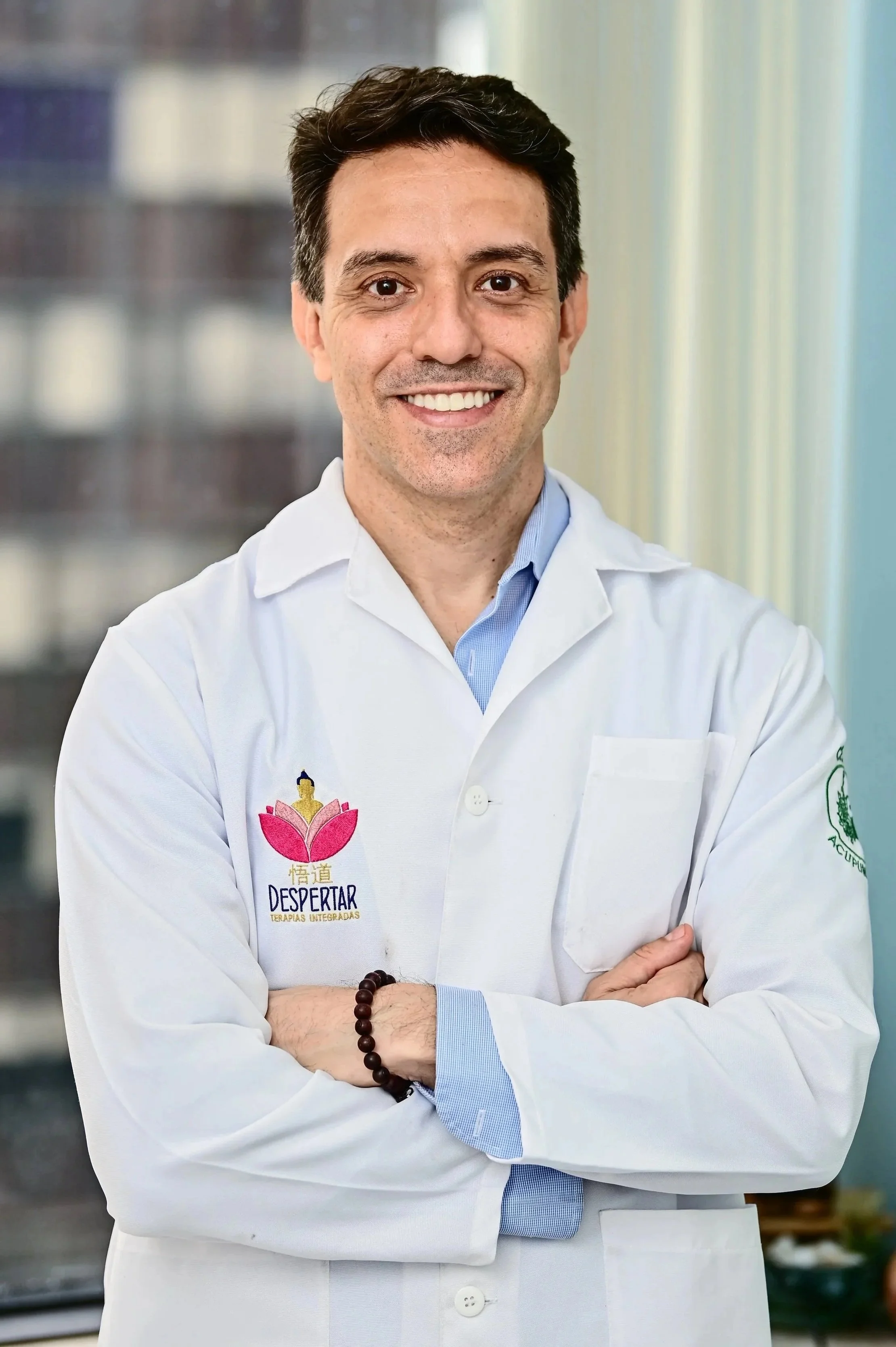 Homem sorridente com cabelo escuro, usando jaleco branco com logotipo de uma flor de lótus rosa e palavras "Despertar Terapias Integradas" na camiseta, de braços cruzados com uma pulseira de contas na mão esquerda, em ambiente interno bem iluminado.