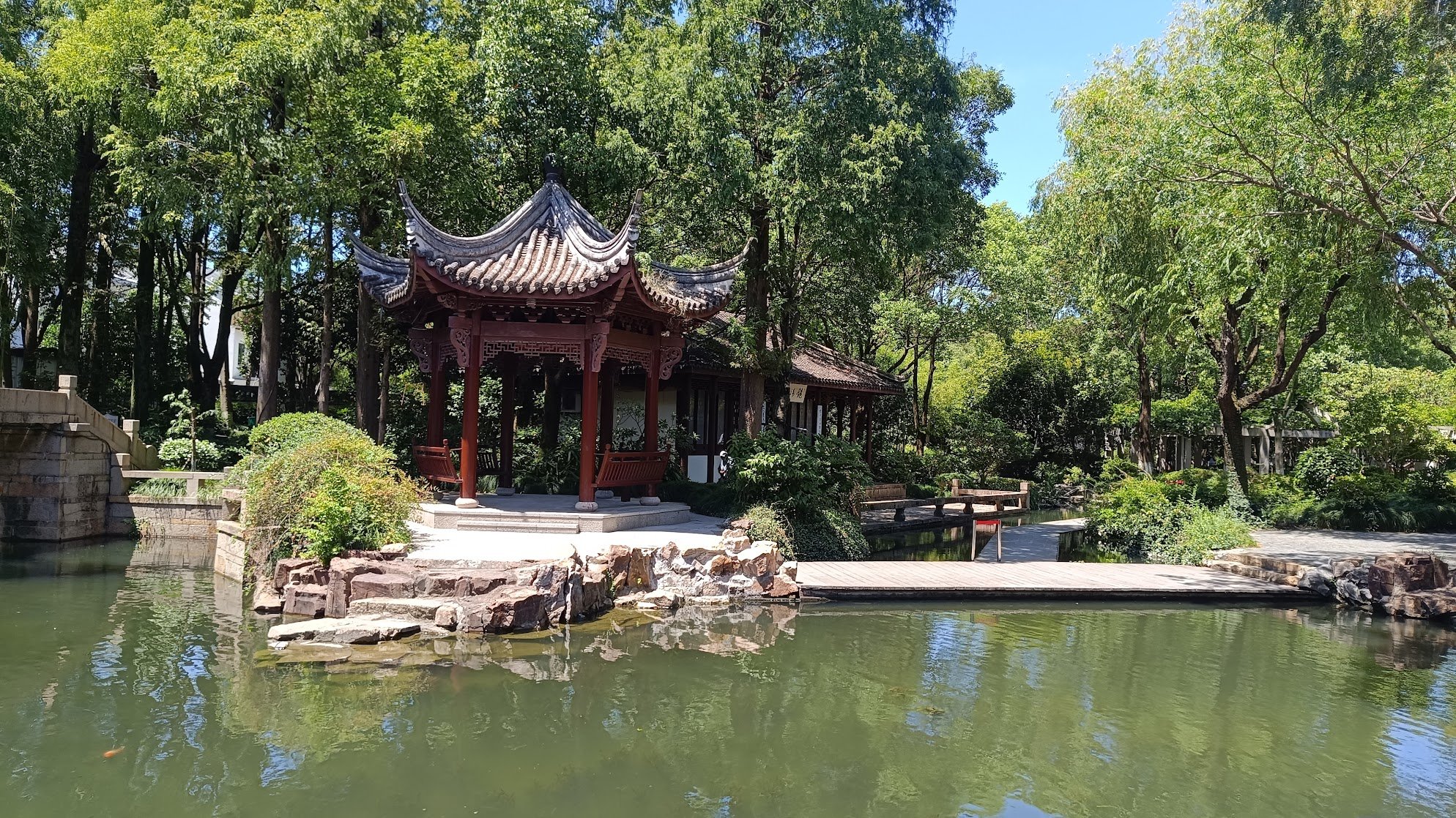 Pavilhão tradicional chinês de madeira com telhado curvado, situado em um jardim com árvores verdes ao redor, próximo à água.