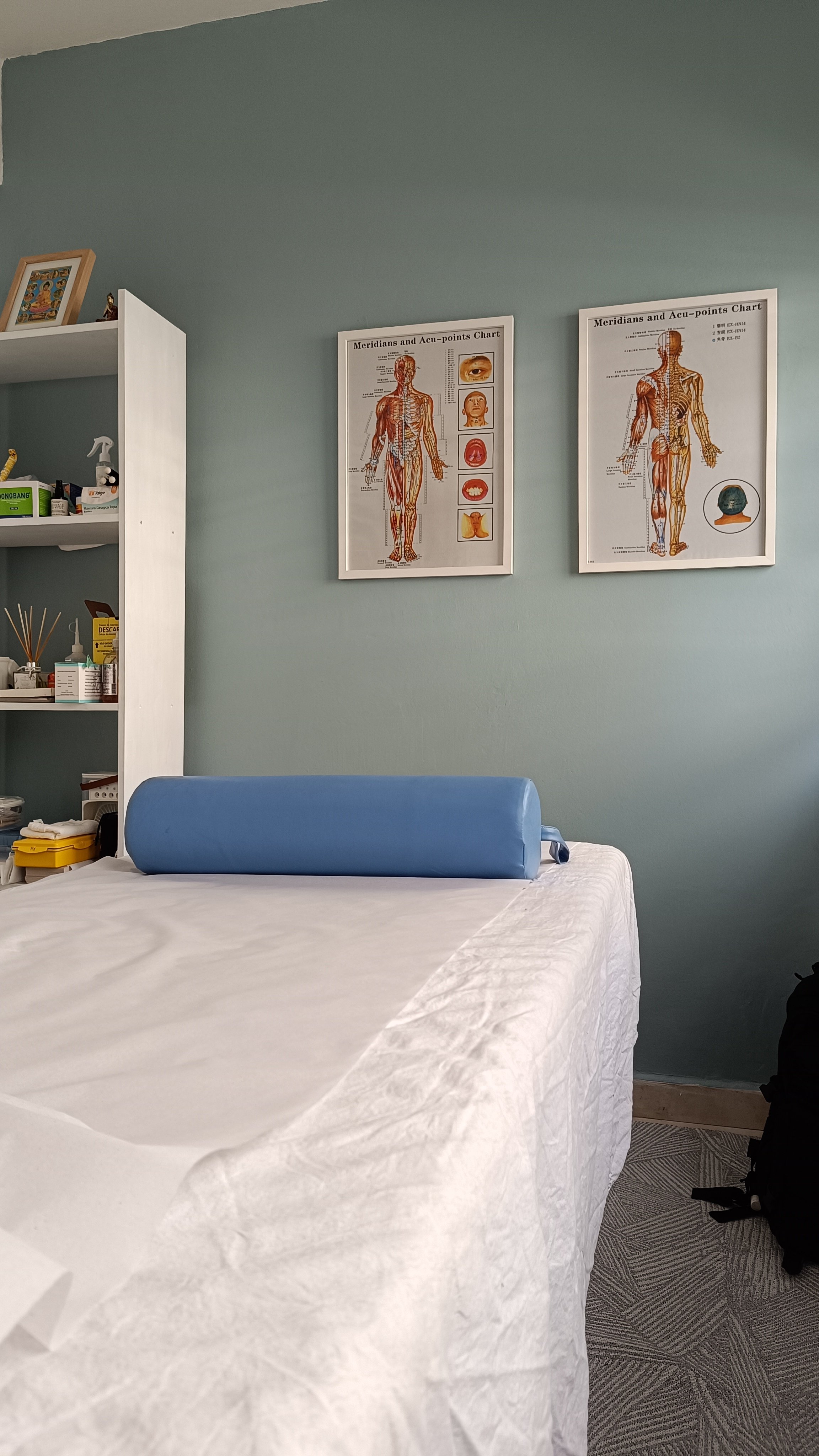 Recurso de tratamento de terapia ou fisioterapia em ambiente clínico, com papel toalha na maca, dois quadros com mapas de pontos de acupuntura na parede, uma estante com produtos de limpeza e medicamentos ao lado.