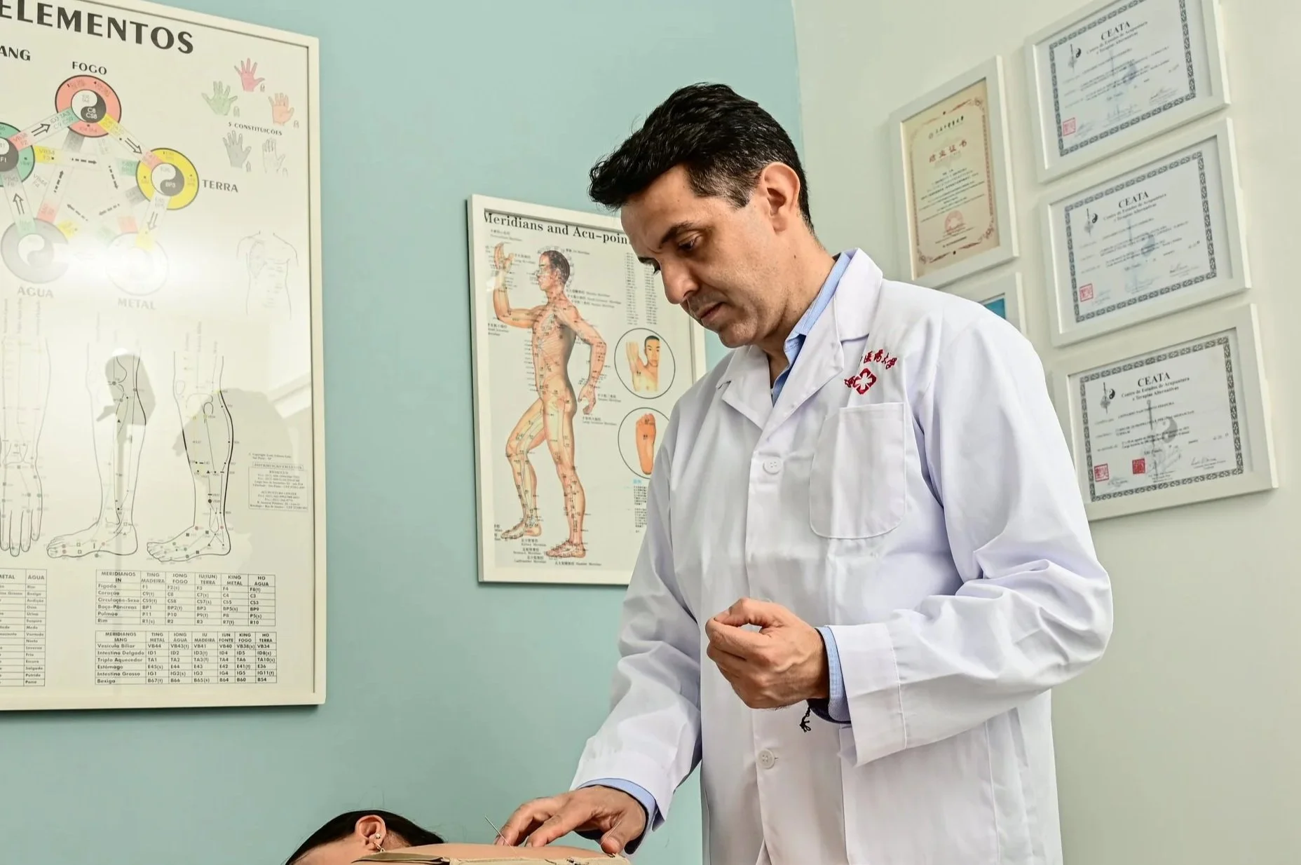Acupunturista realizando tratamento em paciente, com posters de anatomia e pontos de acupuntura nas paredes.