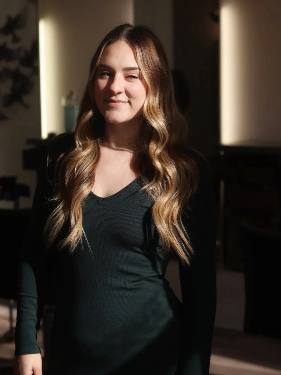 A young woman with long wavy blond hair, wearing a black long-sleeve top, standing in a softly lit room with a slight smile.