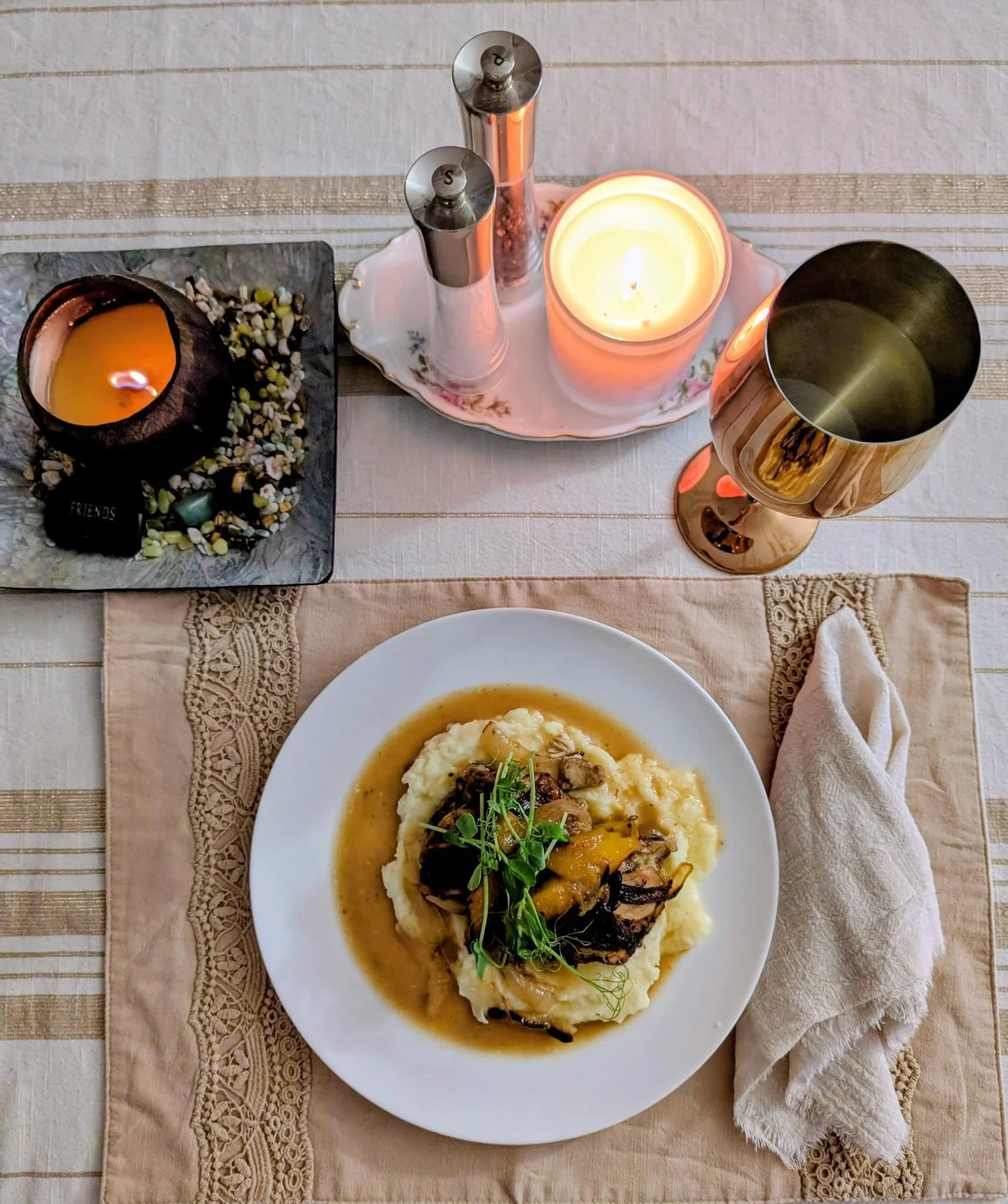 Plate of mashed potatoes with a meat and vegetable topping garnished with microgreens, on a pink placemat. On the table are a lit candle, tall salt and pepper shakers, a rose gold wine glass, and a small black slate with an orange candle and decorative stones.