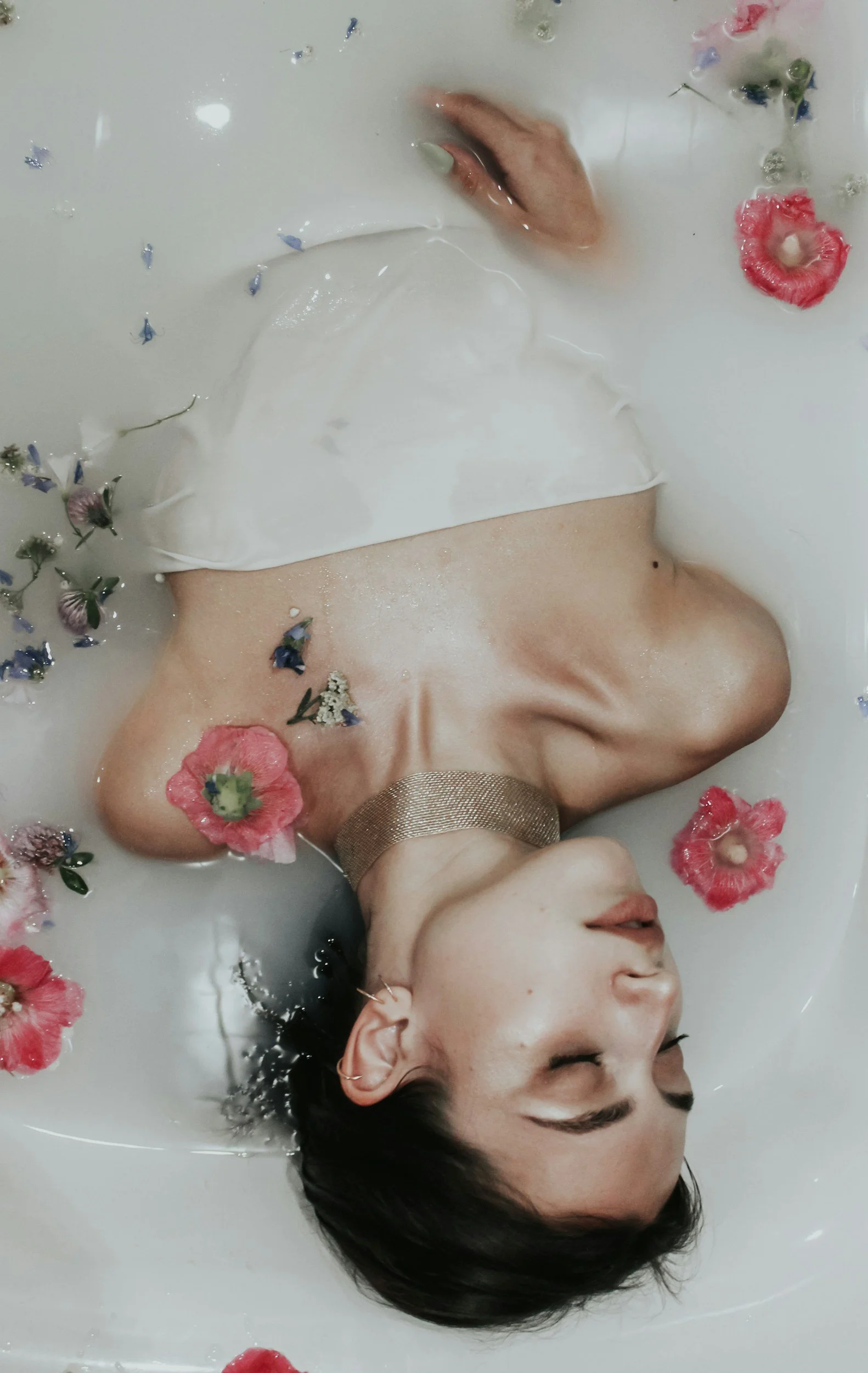 A woman with short dark hair relaxing in a milk bath with floating pink, purple, and white flowers, wearing a white top and a metallic choker.