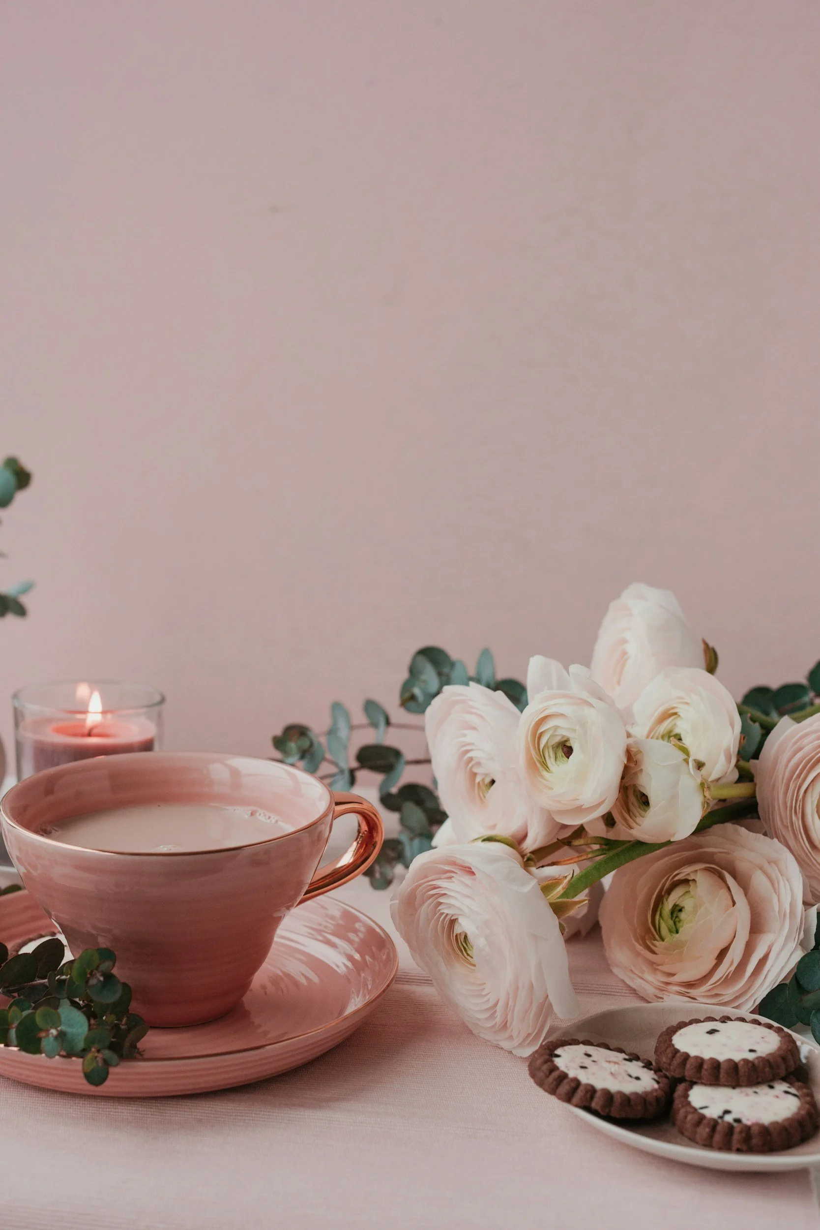 A pink teacup with saucer, white ranunculus flowers, eucalyptus leaves, a lit pink candle in a glass holder, and a plate of decorated cookies on a pink tablecloth against a light pink wall.