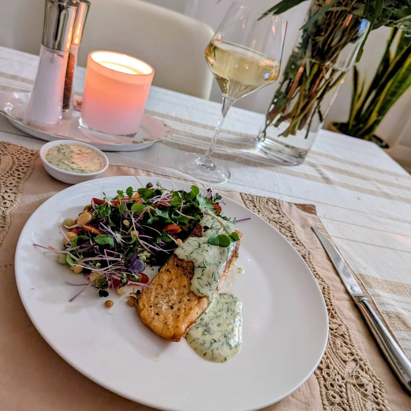A dinner plate with grilled salmon topped with herb sauce, served with a side salad of microgreens and vegetables, on a dining table with a glass of white wine, a candle, and a small bowl of herb sauce. There is a knife placed to the right of the plate.