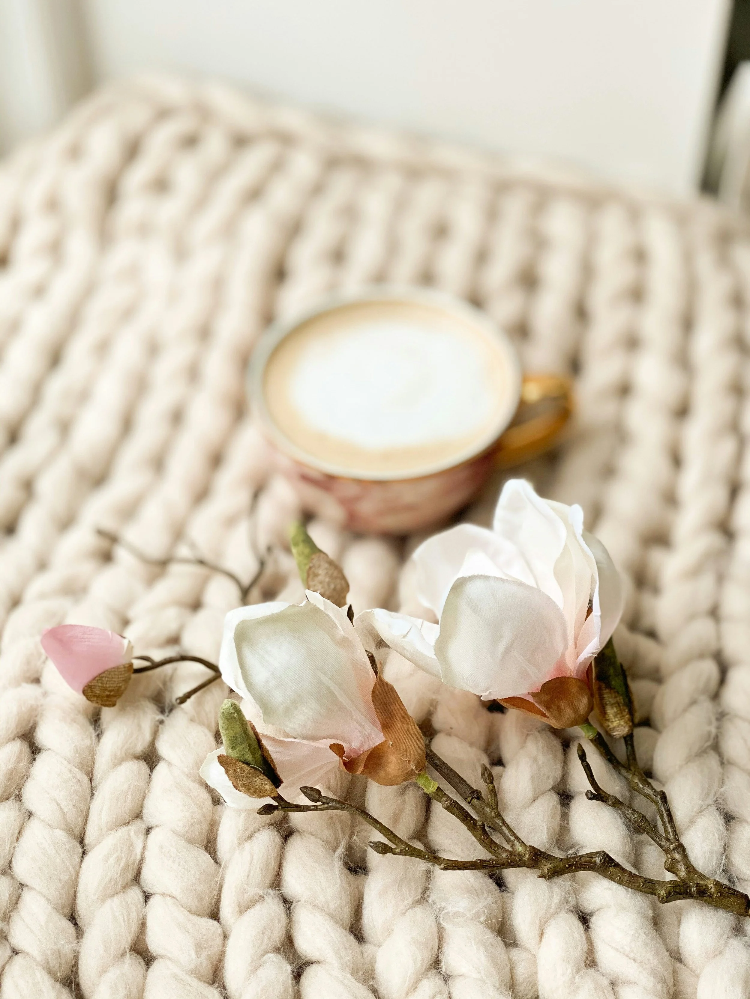 A cup of coffee on a cozy knitted blanket with delicate pink and white magnolia flowers nearby.