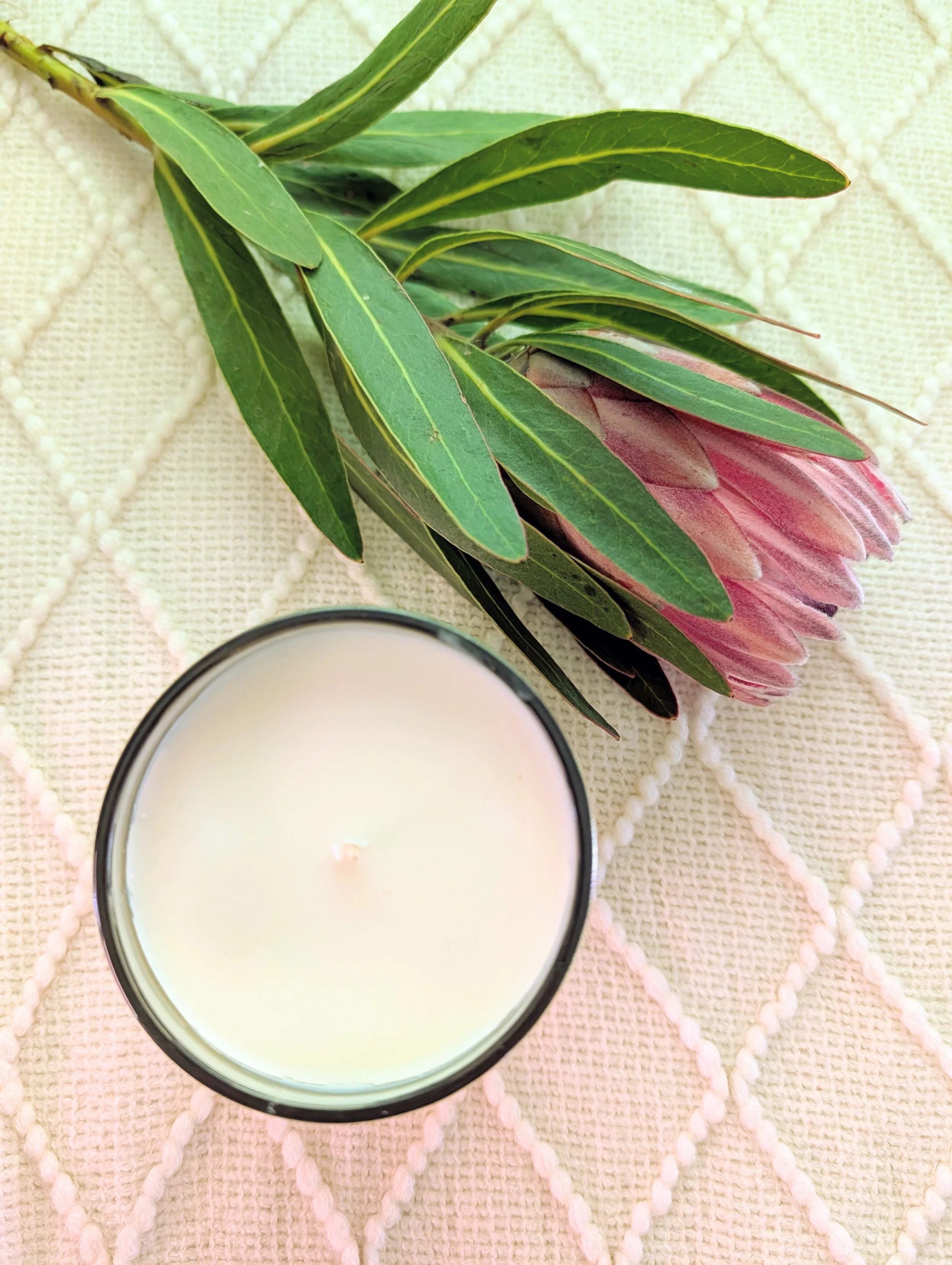 A pink protea flower with green leaves placed next to a candle on a textured surface.