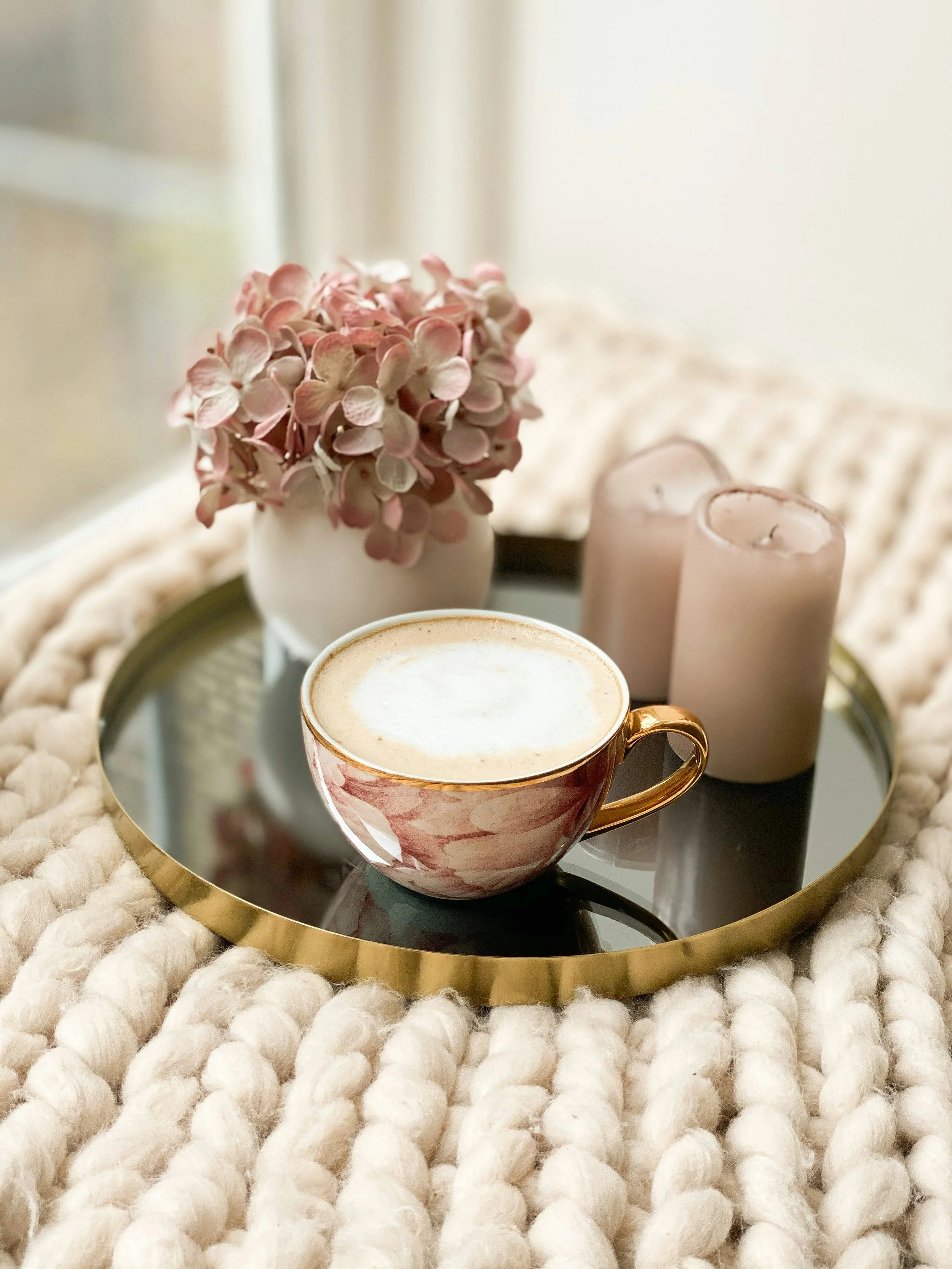 A cozy tabletop arrangement with a floral cup of coffee, pink candles, and a vase of pink hydrangeas on a round gold tray, set on a chunky knitted blanket.
