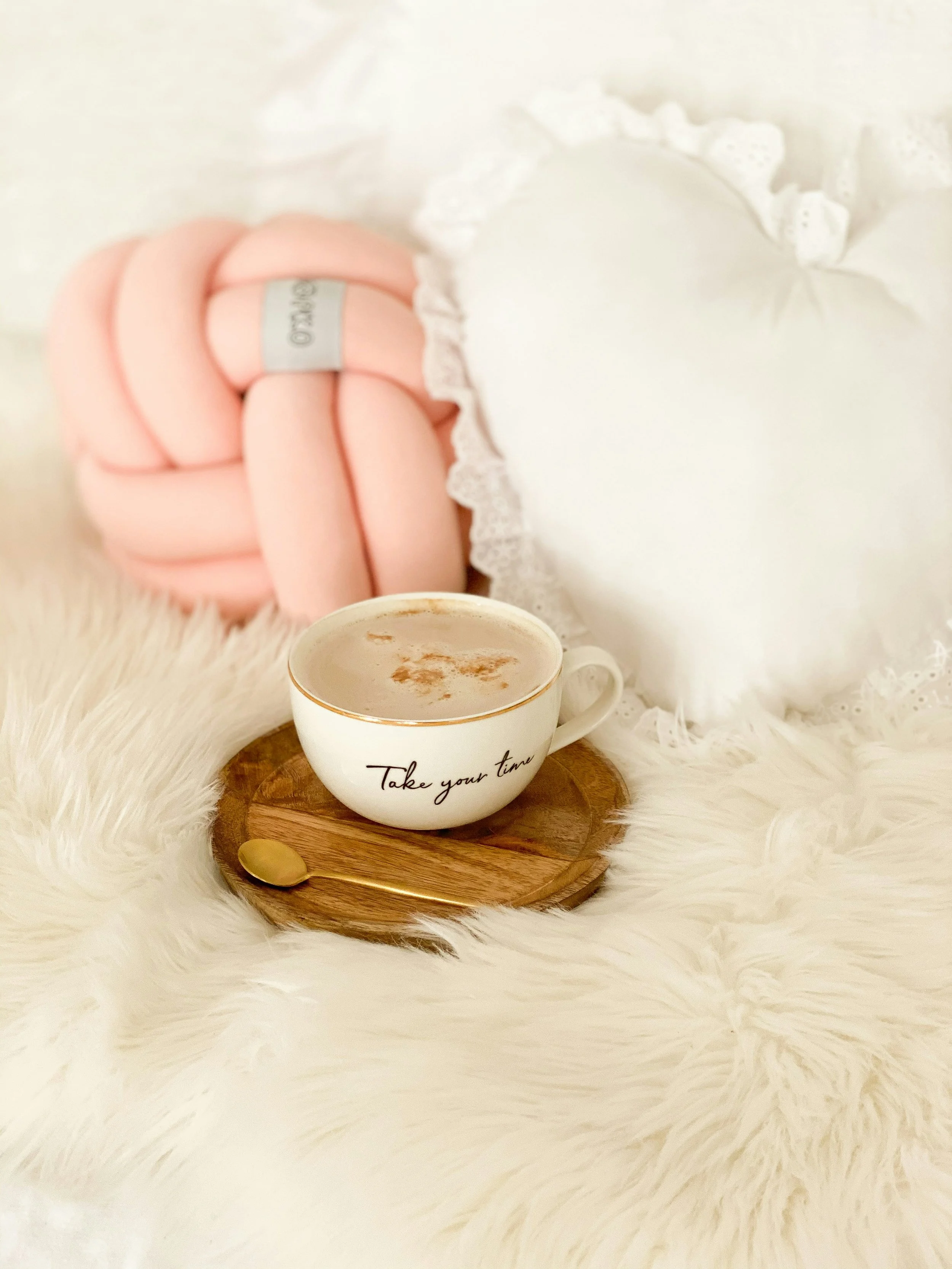 White coffee cup with gold rim and the words "Take your time" written on it, filled with coffee, placed on a small round wooden tray with a gold spoon, surrounded by a white furry surface, with pink marshmallow-like candies and a heart-shaped pillow in the background.