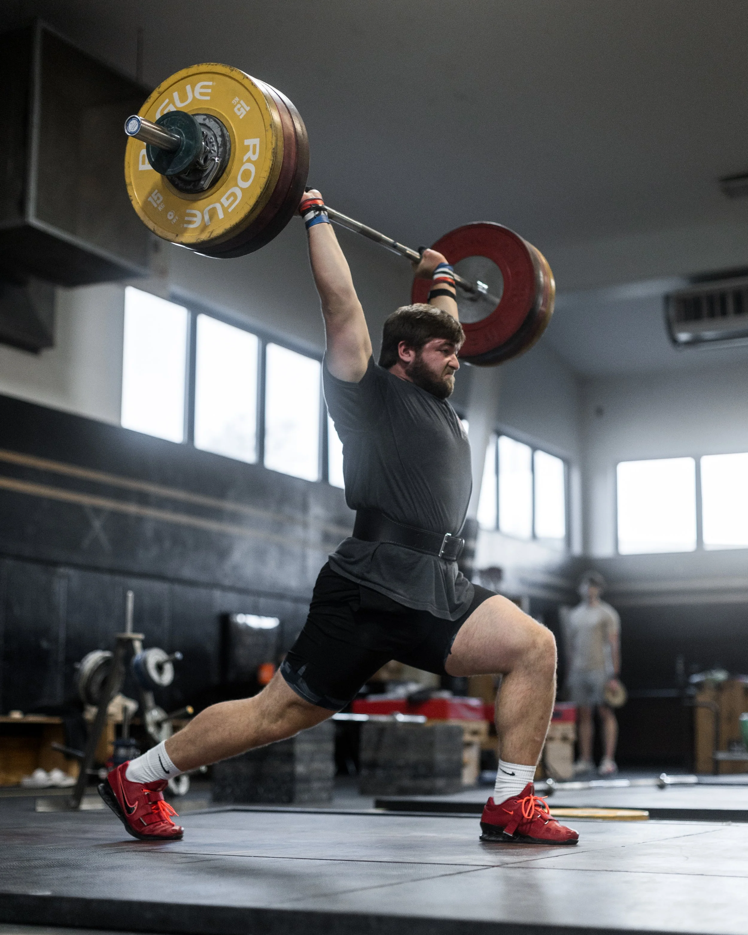 A man in a black t-shirt and shorts is lifting a barbell overhead during a weightlifting workout in a gym, with large windows and a person in the background.