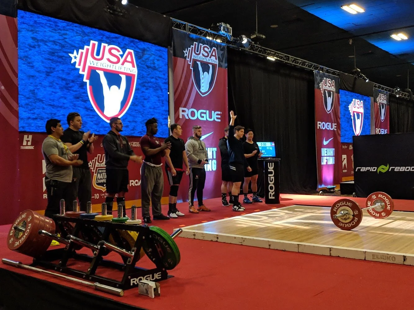 Weightlifting competition stage at USA Weightlifting event with athletes standing on stage, large digital screen displaying USA Weightlifting logo, and weightlifting equipment on the floor.