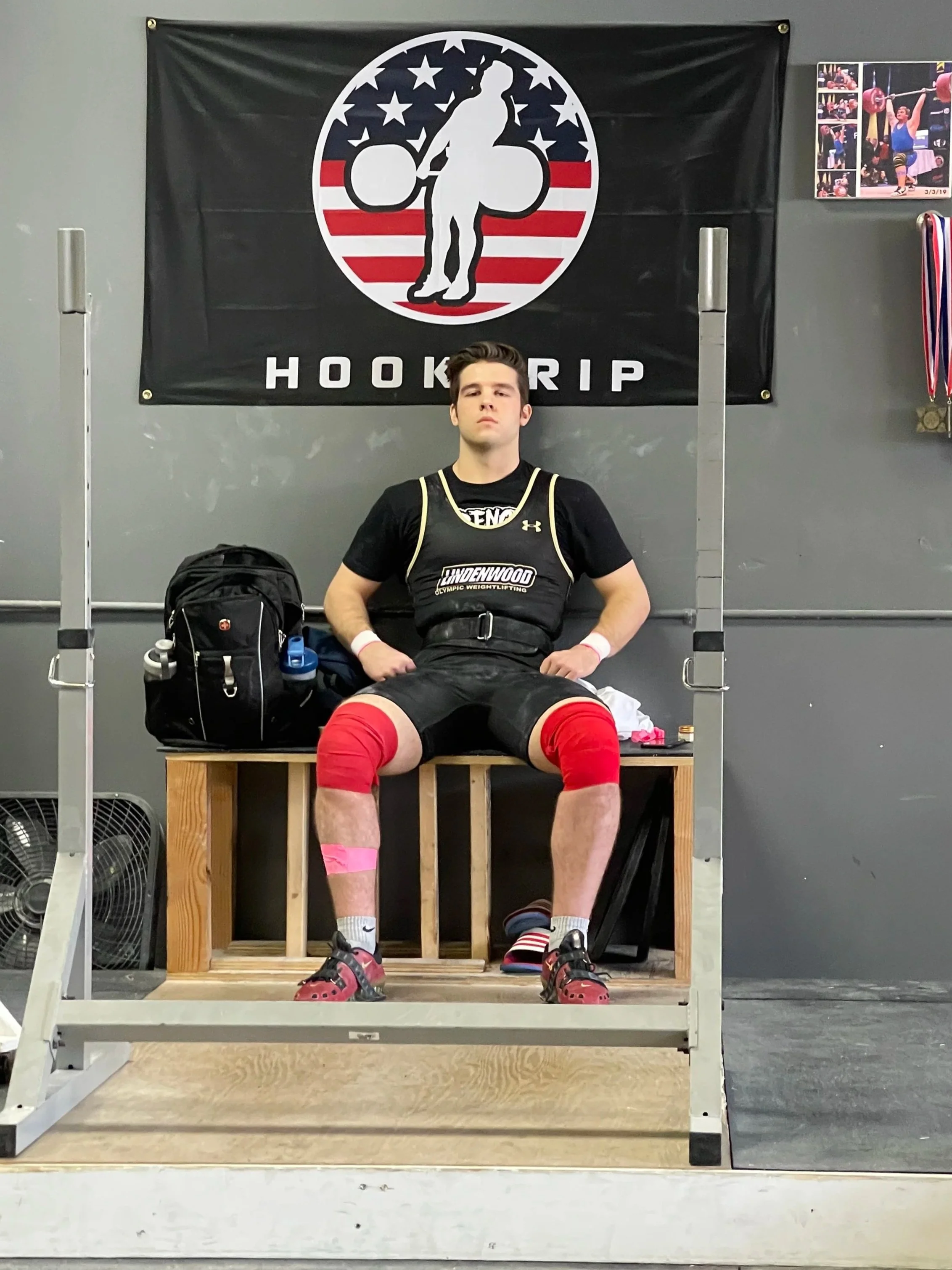 Young man sitting on a wooden bench wearing a weightlifting suit and knee wraps, with a weightlifting gym backdrop and a banner that says 'Hook Grip' behind him.