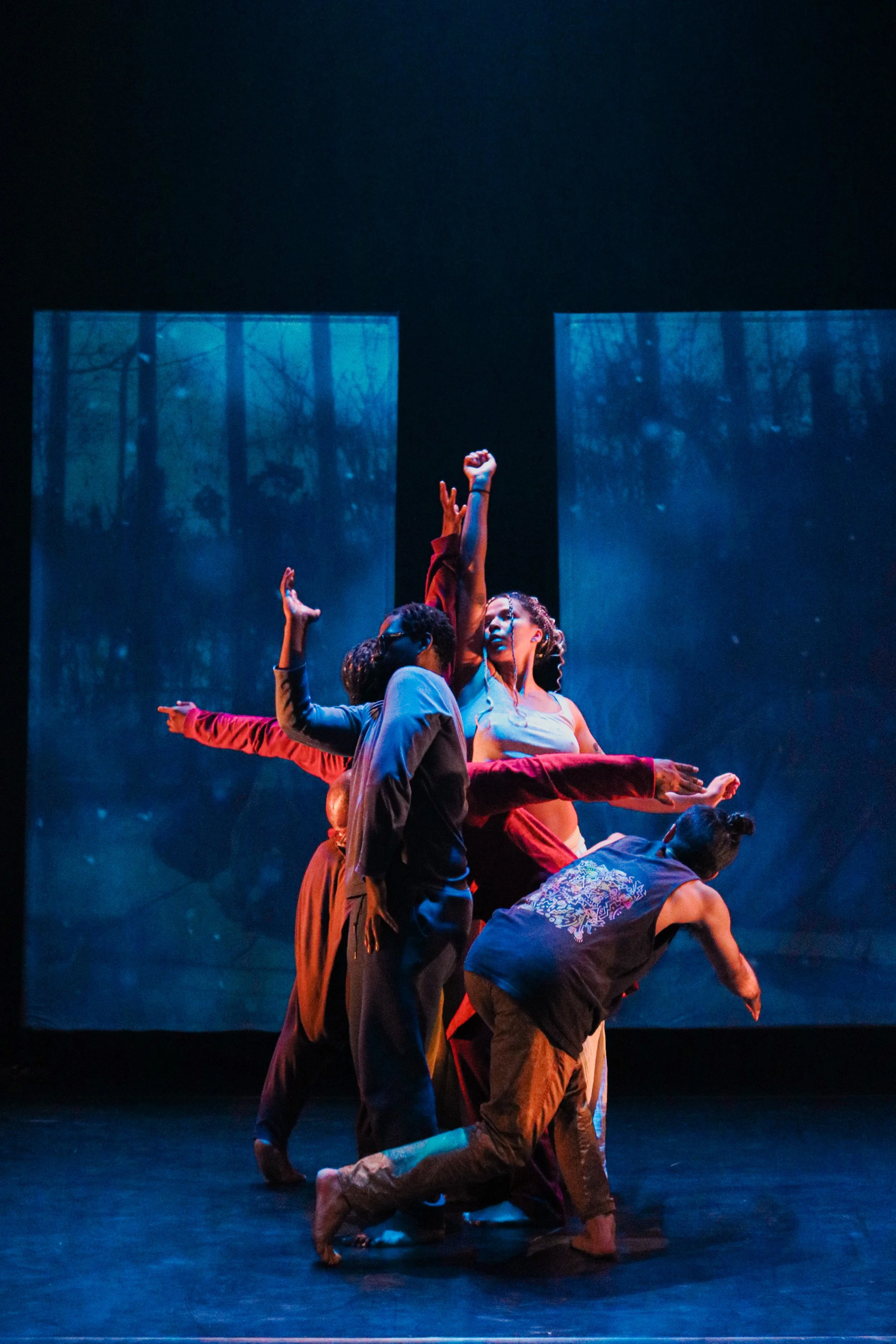 Group of dancers performing on stage with a backdrop of blue-tinted trees, some dancers are standing with arms raised, others are crouched or kneeling.