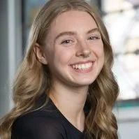 A young woman with long blonde hair smiling against a background of blurred indoor and outdoor elements.
