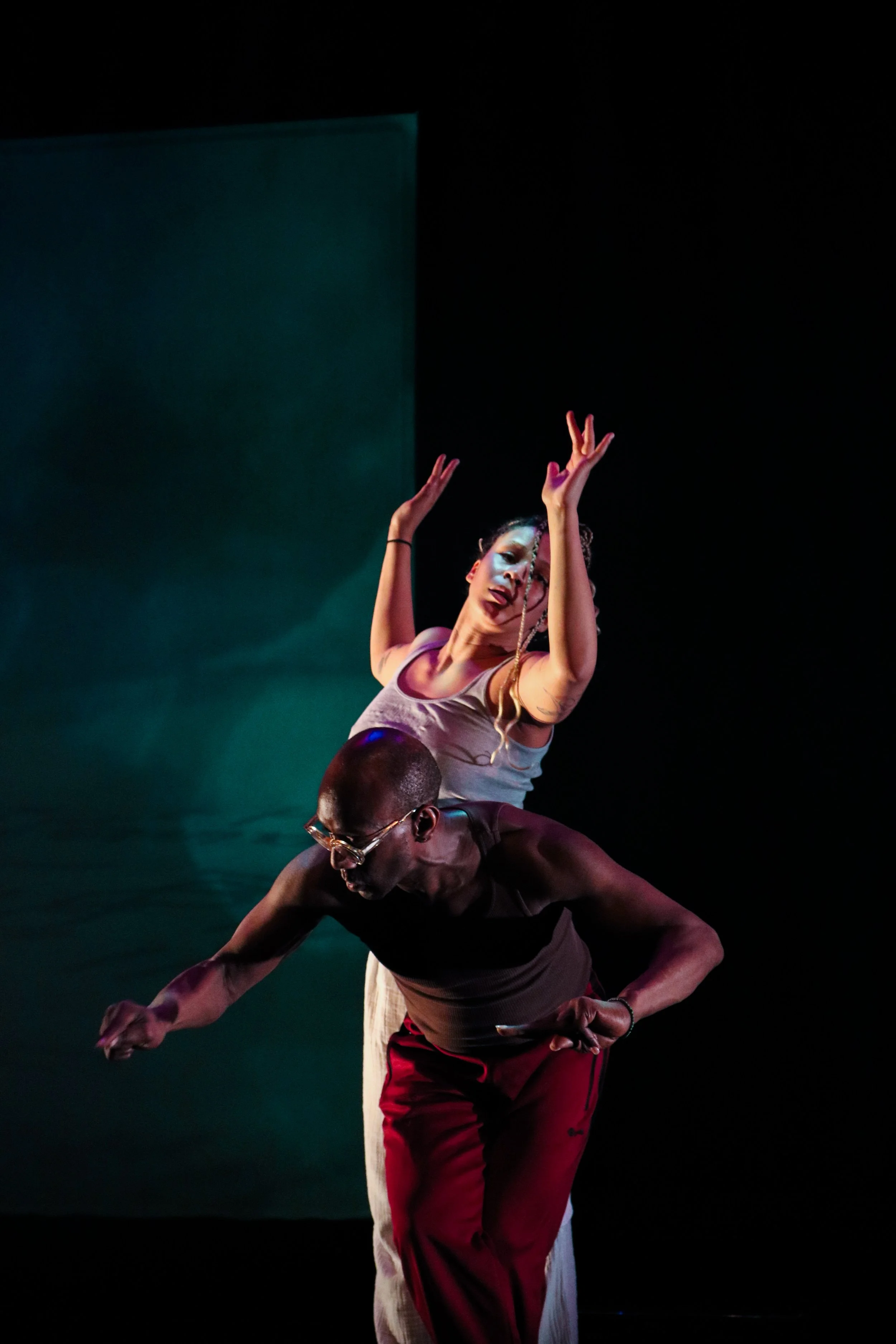 Two performers are on stage, one standing with arms raised and the other leaning forward with a bent posture, both engaged in a dance performance, set against a dark background with teal lighting.