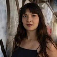 Young woman with long dark hair and bangs, wearing a black sleeveless top, standing outdoors with a tree background.