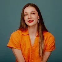 Young woman with long brown hair, wearing an orange shirt, smiling against a teal background.