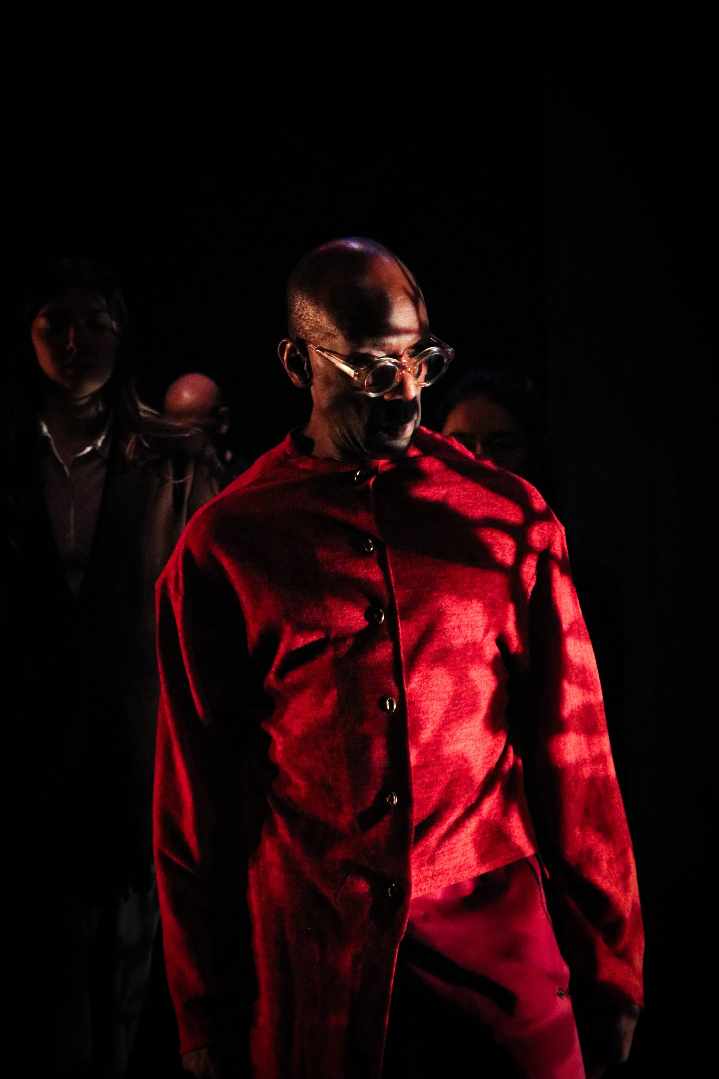 A person with a shaved head and round glasses, wearing a red shirt, standing in a dark setting with dramatic lighting. Three other people are partially visible behind them.