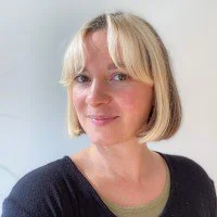 Close-up of a woman with short blonde hair, smiling, against a plain background.