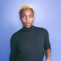 Portrait of a woman with short blonde hair, wearing a black turtleneck, standing against a blue background.