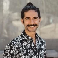A man with dark hair, a mustache, and a goatee, smiling, wearing a black and white patterned shirt, outdoors in a natural setting.