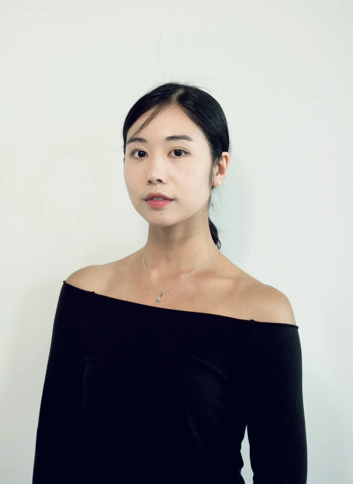 Portrait of a young woman with black hair, wearing a black off-shoulder top and a silver necklace, standing against a plain white background.