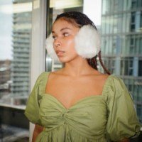 Woman wearing green dress and white earmuffs, standing indoors near large windows.