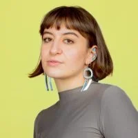 Portrait of a woman with short brown hair and bangs, wearing a gray top and colorful earrings, against a yellow background.