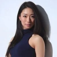 Portrait of a woman with long dark hair wearing a sleeveless blue top, standing against a plain background.