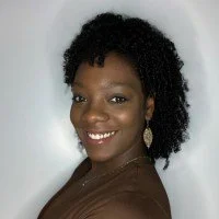 Portrait of a smiling woman with curly black hair, wearing earrings and a brown top against a plain background.