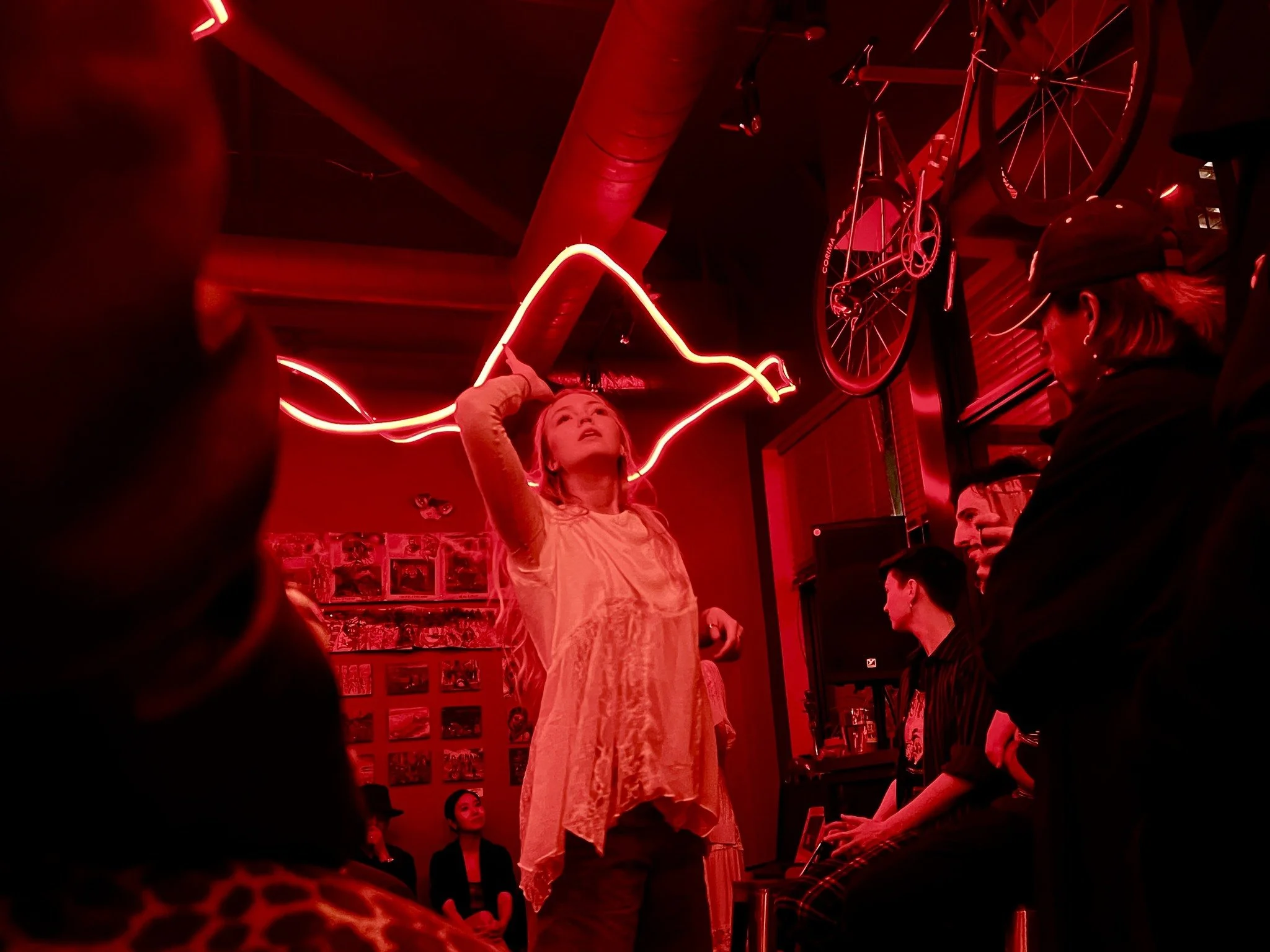 A woman in a white dress performing in a room with red neon lights, surrounded by other people, with a bicycle mounted on the wall above her.