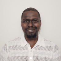 Portrait of a man with dark skin, short hair, and a beard, wearing a light-colored patterned shirt, standing against a light background.