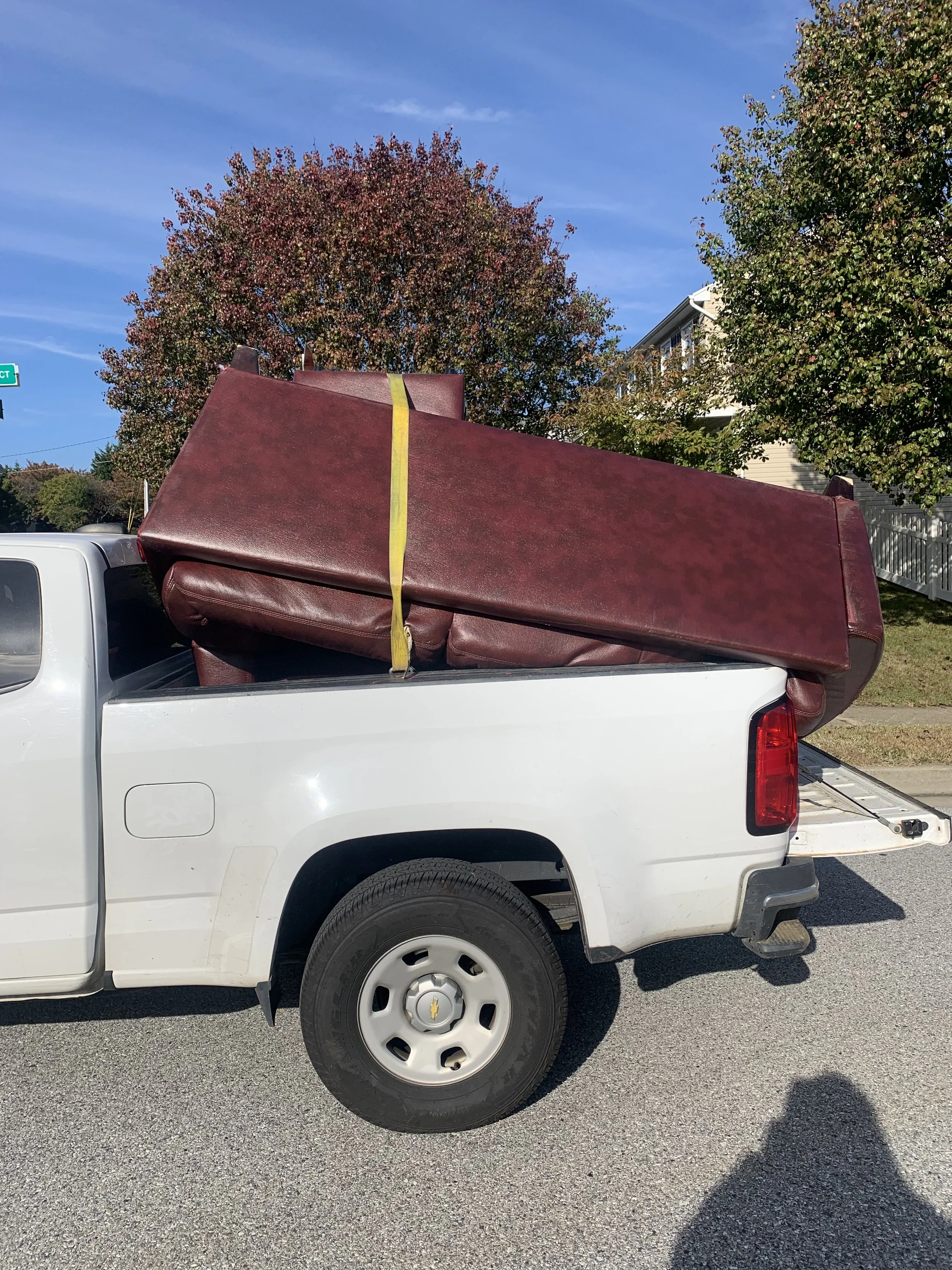 A white pickup truck with a maroon couch loaded in the truck bed, secured with a yellow strap, parked on a residential street with trees and houses in the background.