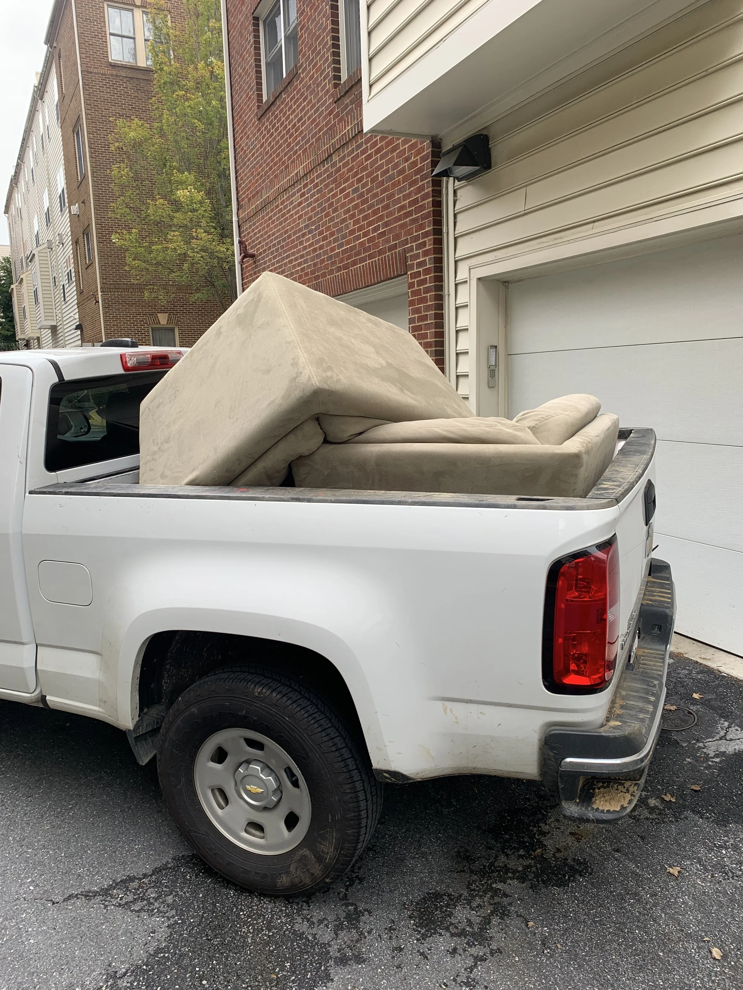 White pickup truck parked in front of a building, filled with large pieces of furniture including a beige sofa and a mattress.