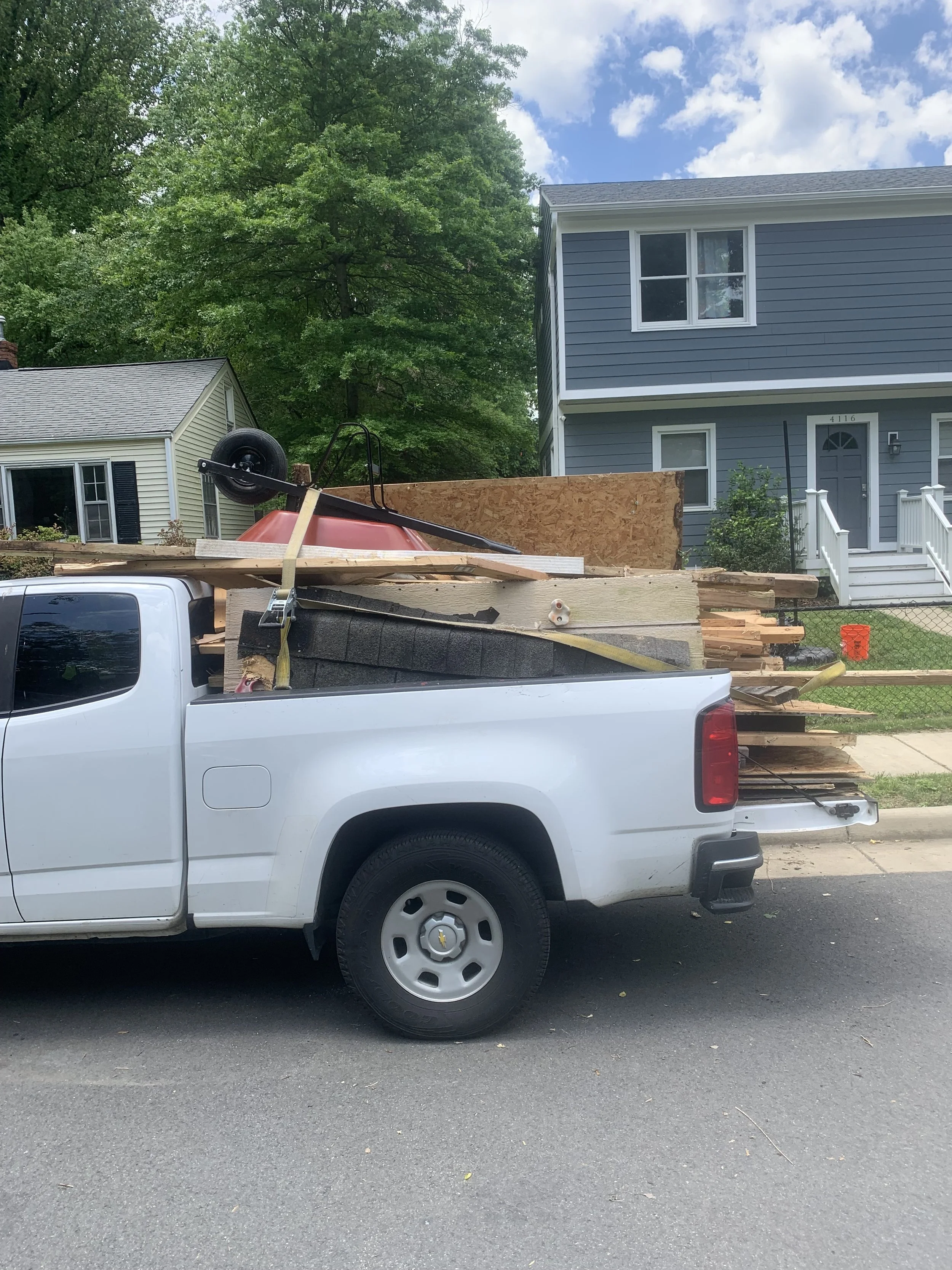 White pickup truck parked on the street loaded with construction materials including lumber, plywood, foam insulation, a wheelbarrow, and other tools, with a blue house and green trees in the background.