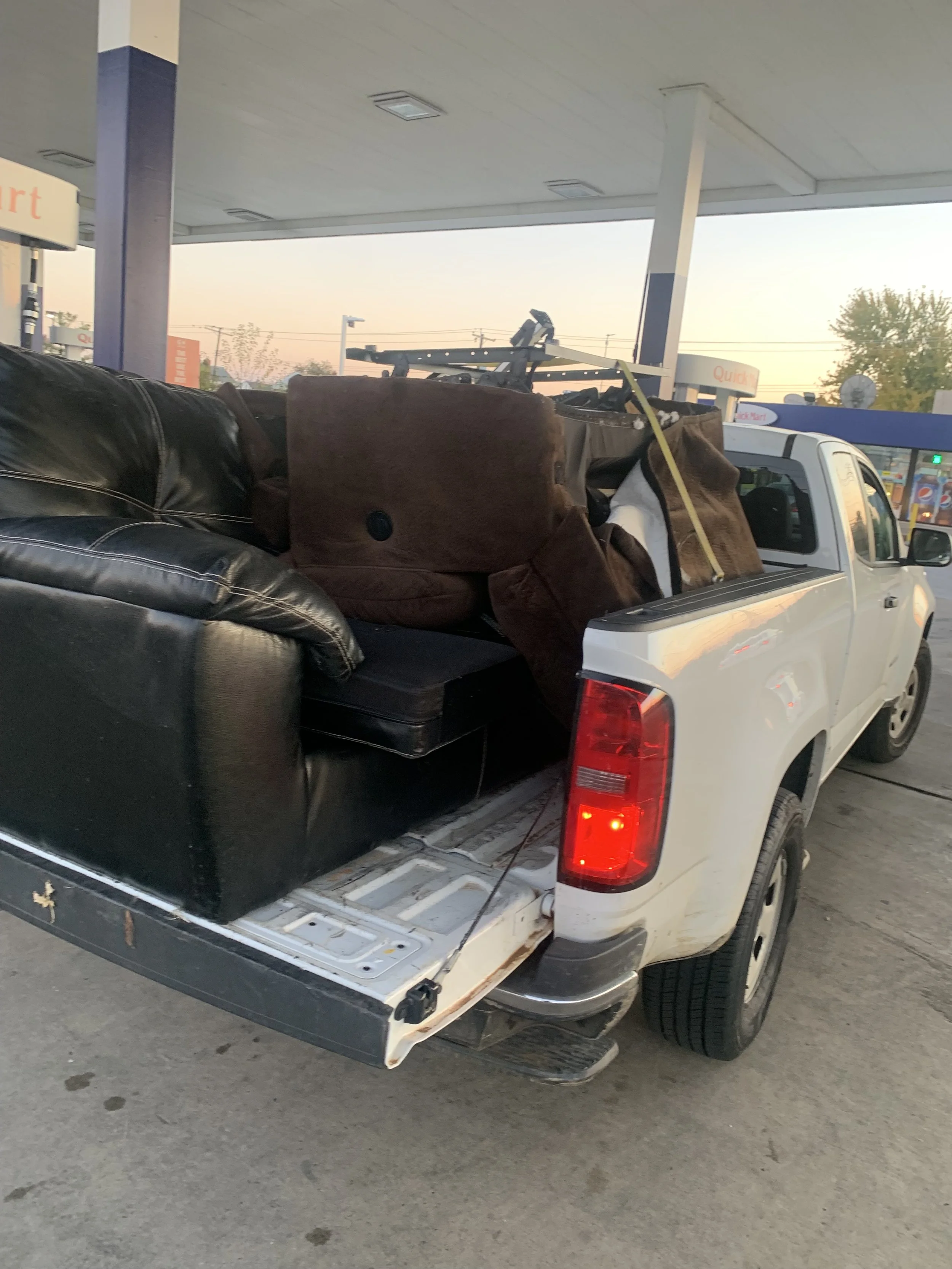 Pickup truck parked at a gas station with furniture loaded in the bed, including a black leather sofa and brown chairs.