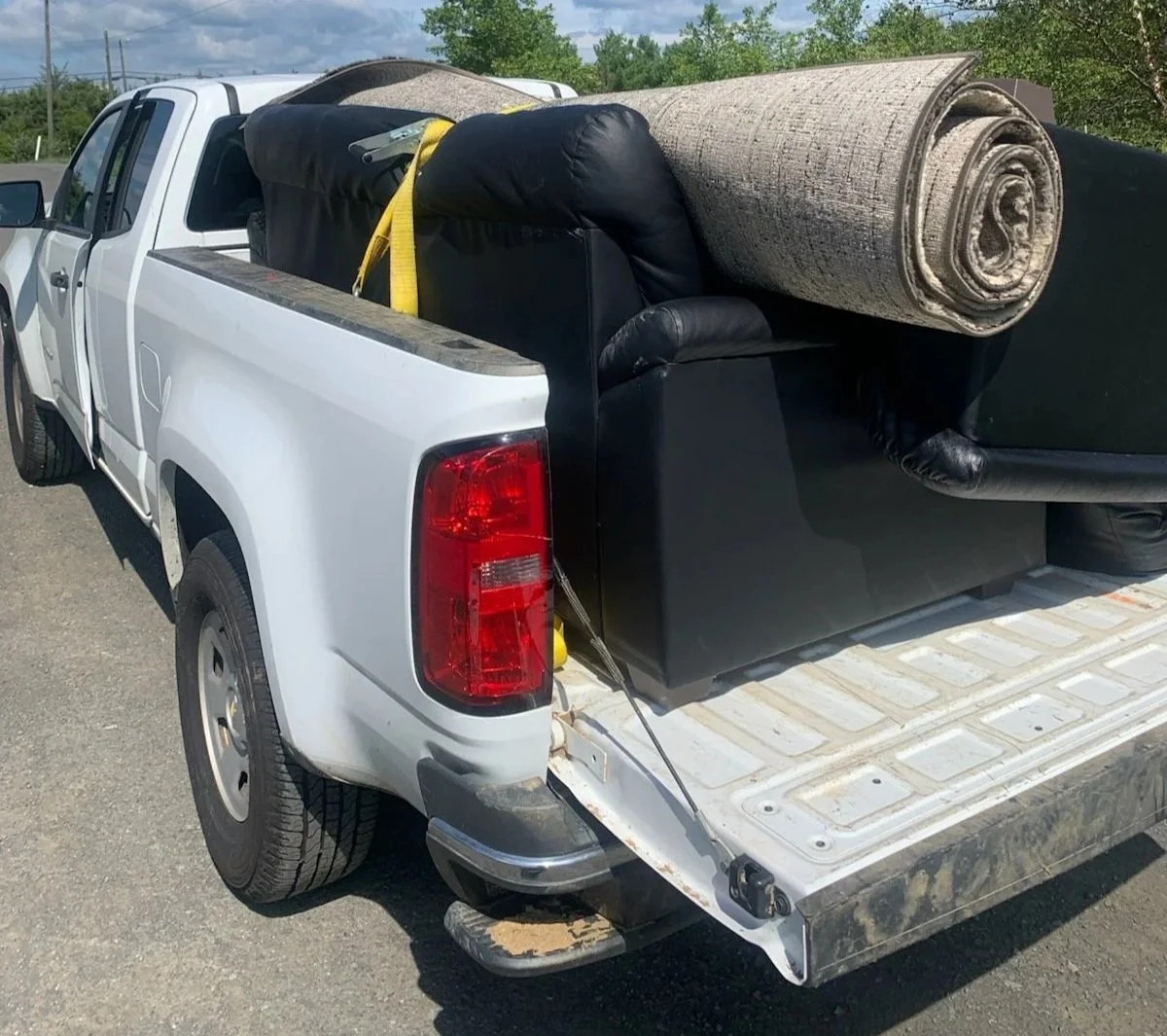 A white pickup truck with tools and furniture in the truck bed, including a rolled-up carpet and a black bag, on a paved surface with green trees in the background.
