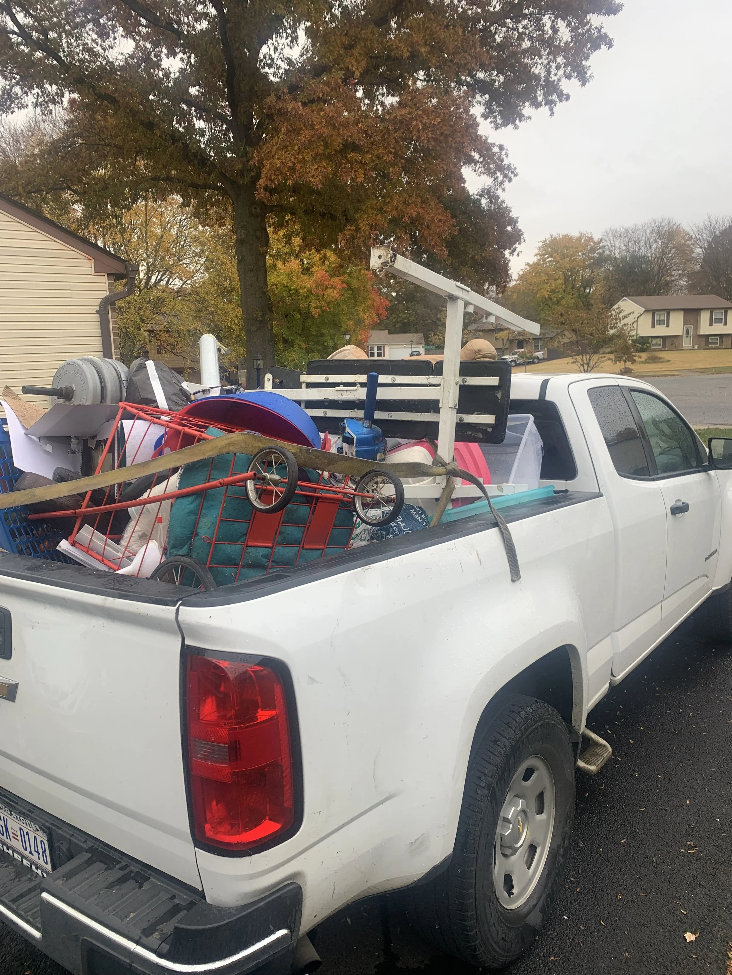 White pickup truck filled with various household items, including a small red shopping cart, a blue item, a white wooden frame, and other miscellaneous objects, parked outdoors on a rainy day.