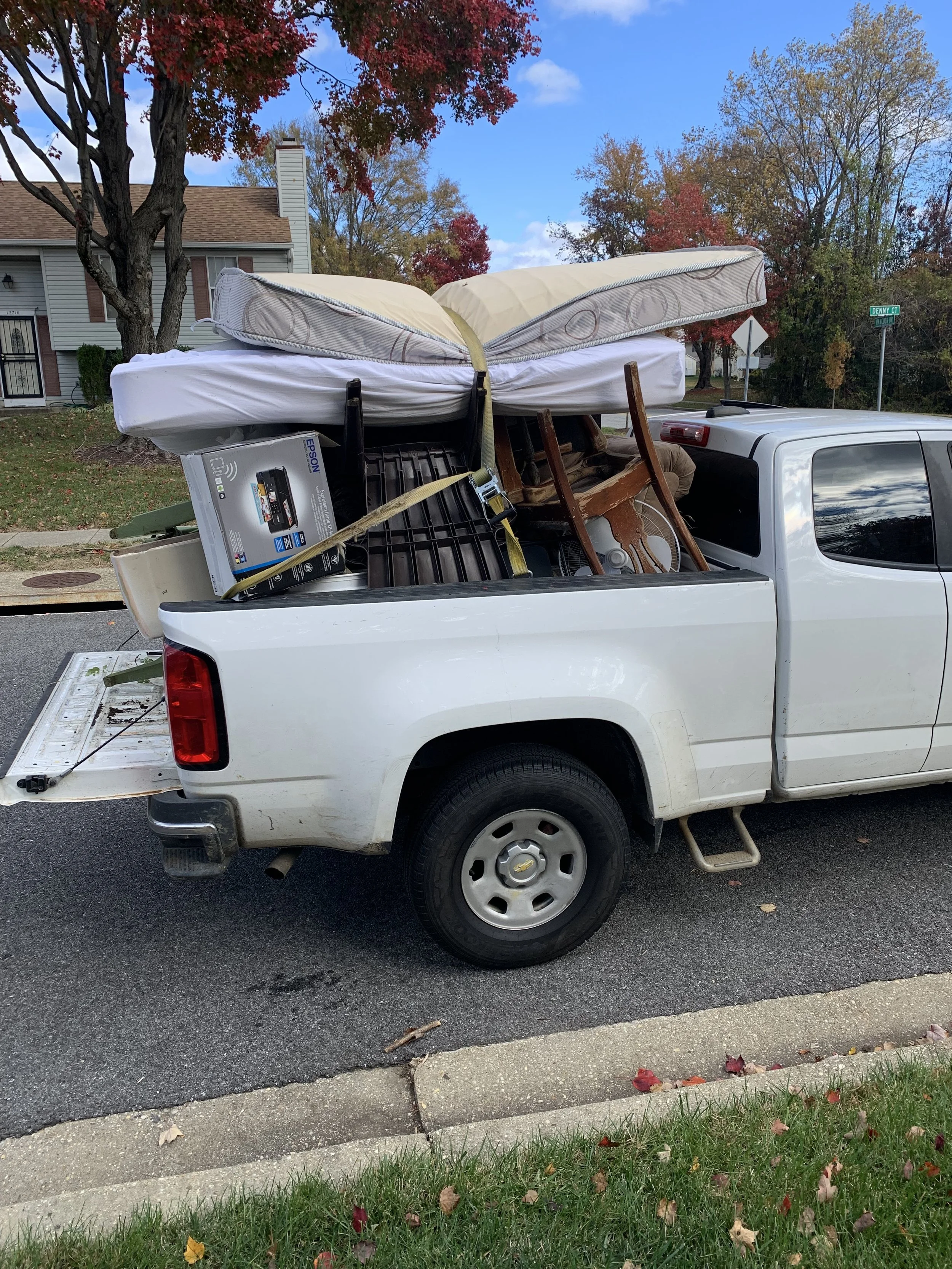White pickup truck parked on the street loaded with furniture and household items, including a mattress, chairs, and a boxed printer.