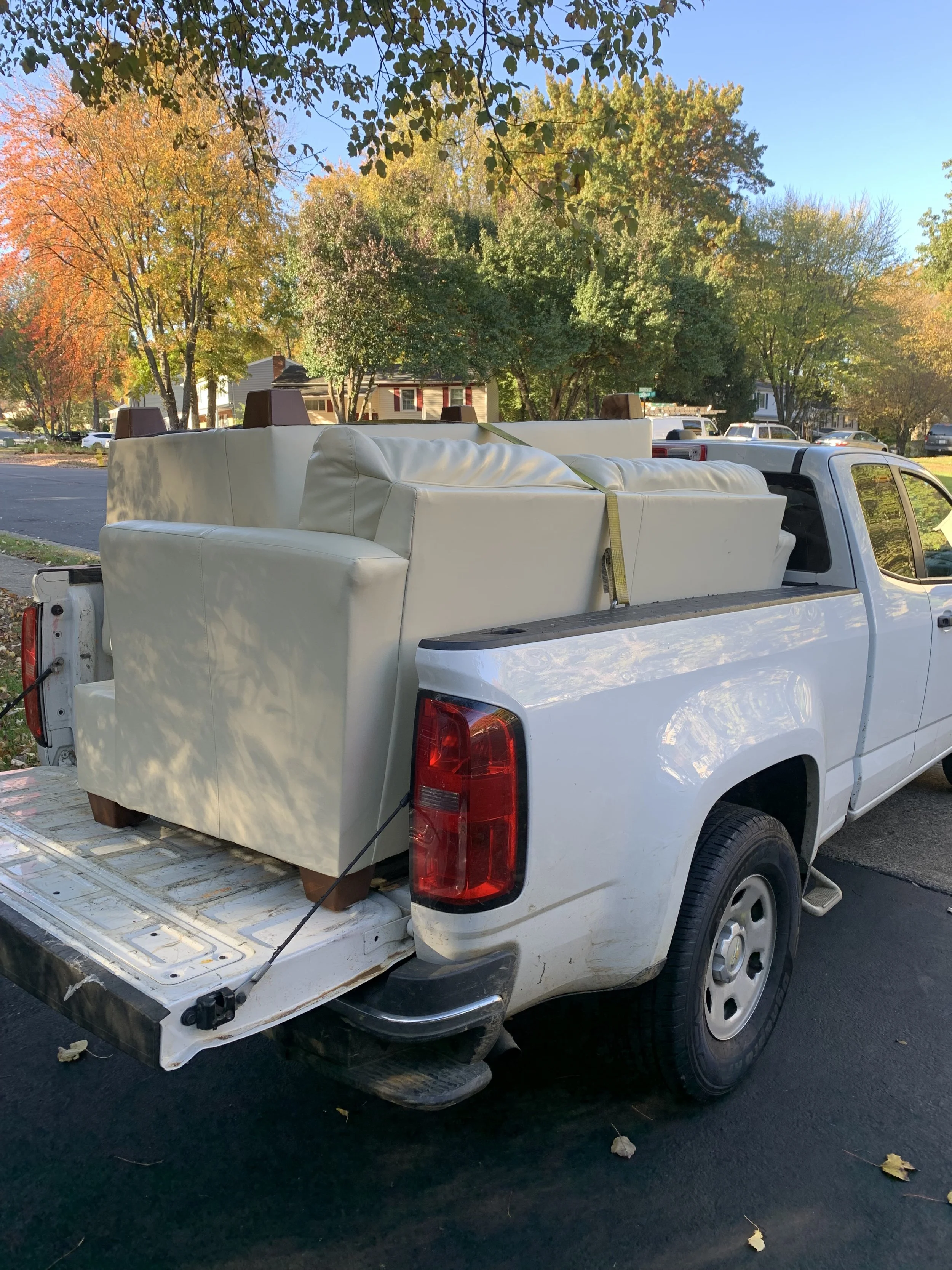 A white pickup truck parked in a residential neighborhood with autumn foliage. The truck's bed contains large white furniture pieces, including sofas, secured with straps.
